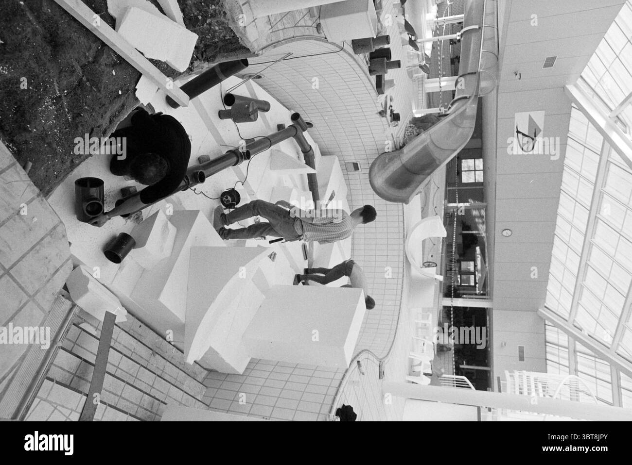 Lavorare sulla diapositiva Gran Dorado, Whizgle News, Dutch Desk, Paesi Bassi, 1950 - 2000 il 09-02-1992. L'immagine contiene questi argomenti. La scena raffigura un cantiere o un cantiere di installazione in interni, caratterizzato da un ampio spazio aperto illuminato da luce naturale diffusa e luminosa che filtra attraverso un soffitto in vetro. L'ambiente sembra essere un edificio moderno con linee pulite e un'estetica contemporanea. In primo piano, due persone sono attivamente impegnate nel loro lavoro. Una persona, posizionata a sinistra, è accovacciata, concentrandosi su un contenitore mentre applica un liquido più scuro Foto Stock