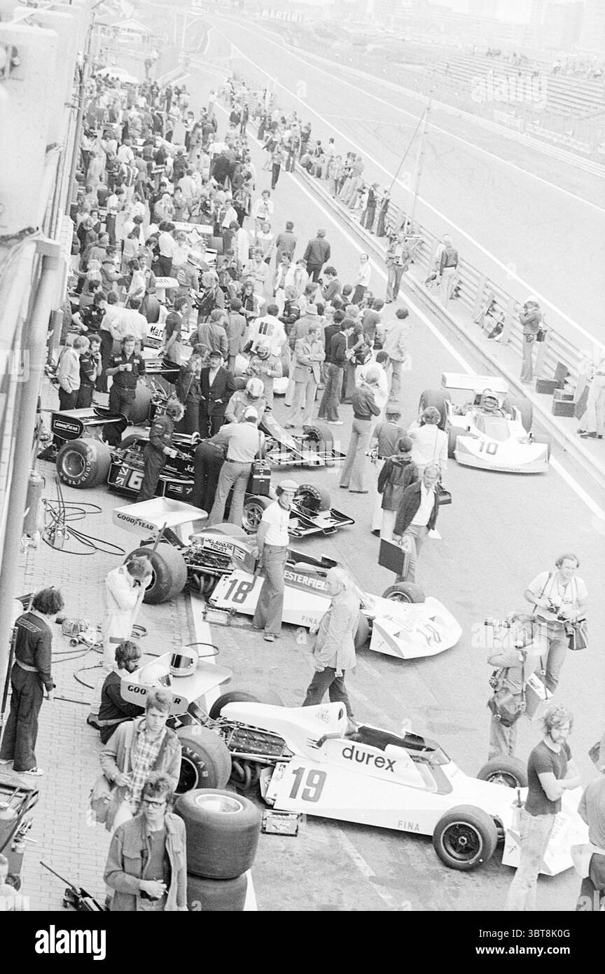 Gare automobilistiche Grand Prix, Whizgle News, Dutch Desk, Paesi Bassi, 1950 - 2000 on 27-08-1976. L'immagine contiene questi argomenti. La scena raffigura un vivace pit Lane per gli sport motoristici, caratterizzato da una miscela di emozioni e anticipazioni. Molte persone popolano l'area, dal personale di squadra agli spettatori, tutti impegnati in varie attività legate all'evento. Sulla sinistra, diverse eleganti auto da corsa, principalmente in bianco e adornate con loghi sponsor, occupano i posti ai box. Il loro design trasuda un senso di velocità e precisione, visibile anche a riposo. Gli pneumatici sono impilati nelle vicinanze, indicando Foto Stock