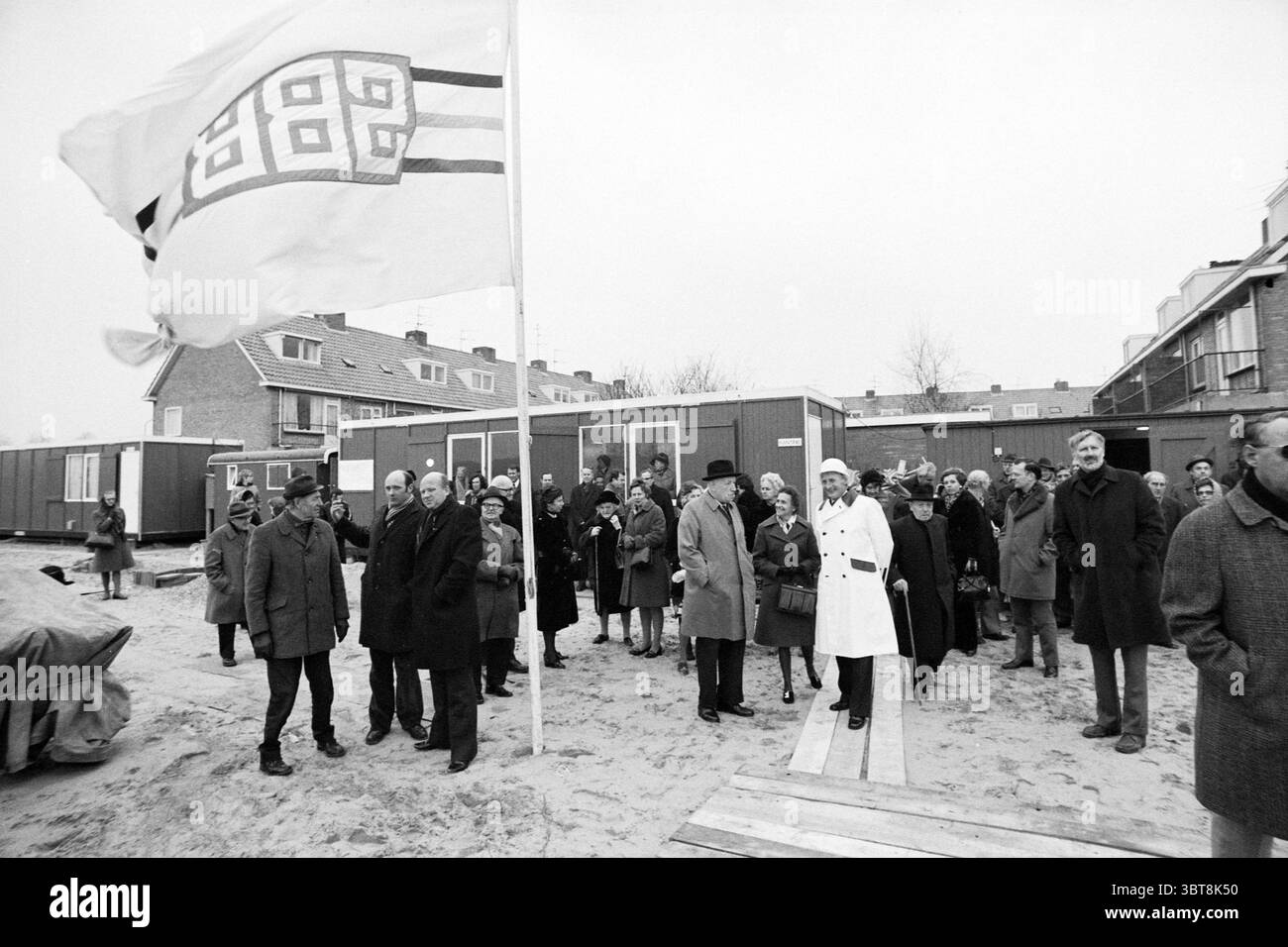 Prima pila di casa di riposo Helmlaan prima pila, Whizgle News, Dutch Desk, Paesi Bassi, 1950 - 2000 il 08-03-1974. L'immagine include questi argomenti. In primo piano, un gruppo diversificato di persone si trova su una superficie sabbiosa, probabilmente un'area all'aperto per eventi. Gli individui sono vestiti con vari strati di abbigliamento, suggerendo un ambiente fresco; molti indossano cappotti, mentre alcuni sport sciarpe e cappelli. La folla sembra impegnata, con espressioni che vanno dalla curiosità alla solennità. Sullo sfondo si possono vedere diverse strutture prefabbricate, possibilmente edifici temporanei o uffici. Il loro mu Foto Stock