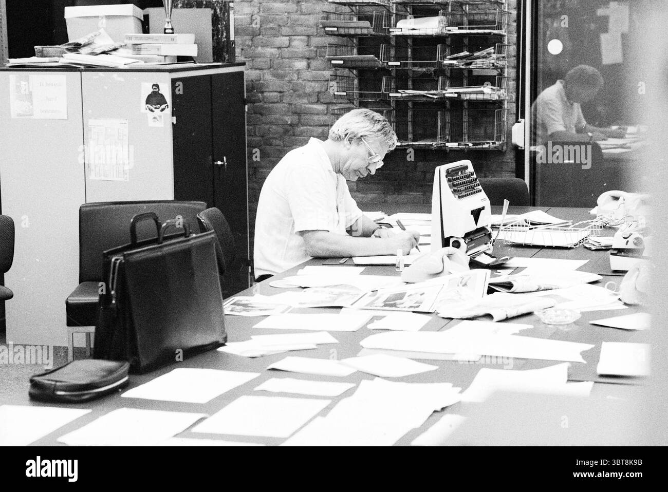 Jan Blijboom direttore sportivo. HD Haarlems Dagblad ecc. e IJmuider Courant e Koeri, Whizgle News, Dutch Desk, Paesi Bassi, 1950 - 2000 il 24-09-1980. Questi argomenti vengono visualizzati nell'immagine. In un ambiente di lavoro vivace, la scena cattura un momento di concentrazione concentrata. Un uomo di mezza età, con capelli e occhiali argentati, si appoggia su una scrivania disordinata, avvolto nella scrittura su un foglio di carta. La sua espressione riflette un pensiero profondo, punteggiato da sopracciglia a solco. Intorno a lui, la scrivania è una caotica serie di documenti, note sparse e vari documenti, che fanno pensare a un ambiente di lavoro affollato. Pile di stampe Foto Stock