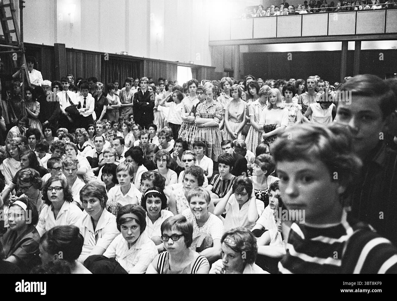 North Holland Philarmonic Orchestra Music, Whizgle News, Dutch Desk, Paesi Bassi, 1950 - 2000 il 09-06-1963. Questi sono gli elementi dell'immagine. La scena raffigura un grande raduno di persone in un ambiente interno, probabilmente durante un evento pubblico o un'assemblea. L'atmosfera è piena di un senso di anticipazione e di coinvolgimento collettivo. In primo piano, un gruppo eterogeneo di individui, prevalentemente giovani, si siedono a stretto contatto sul pavimento, esprimendo un mix di curiosità e concentrazione. Molte persone indossano camicie e abiti in una vasta gamma di modelli classici, alcuni a scacchi, altri a righe Foto Stock