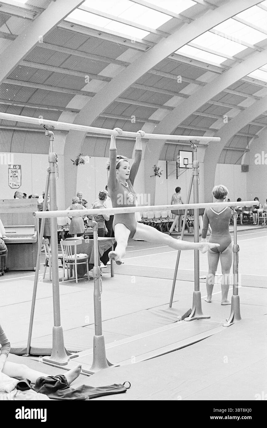 Ginnastica Paesi Bassi - Cecoslovacchia ginnastica, Whizgle News, Dutch Desk, Paesi Bassi, 1950 - 2000 il 03-07-1966. Questi sono gli elementi dell'immagine. La scena si sviluppa in una spaziosa palestra, caratterizzata da un alto soffitto adornato da archi che donano un'atmosfera ariosa. La luce naturale inonda attraverso le grandi finestre, creando un'illuminazione soffusa che migliora l'atmosfera. Le pareti sono fiancheggiate da varie attrezzature da ginnastica e il pavimento è coperto da un morbido tappeto. In primo piano, una giovane ginnasta, rivestita con un cordone aderente alla forma, è aggraziatamente appesa a un insieme di barre parallele. Lei Foto Stock