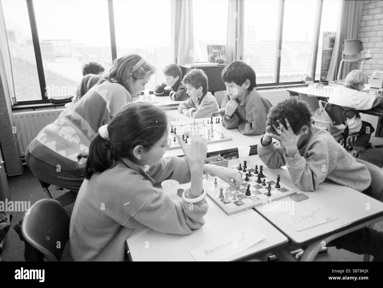 Concorso scolastico di scacchi Bavoschool Chess, Whizgle News, Dutch Desk, Paesi Bassi, 1950 - 2000 il 13-01-1988. Questi argomenti sono mostrati nell'immagine. La scena cattura una classe piena di attività e concentrazione. La luce naturale scorre attraverso le grandi finestre, proiettando ombre soffuse in tutta la sala e creando un'atmosfera calda e invitante. In primo piano, un gruppo di bambini è riunito intorno a diverse scacchiere disposte sulle loro tavole. I pezzi degli scacchi, alternati tra colori scuri e chiari, sono meticolosamente disposti, alcuni a metà partita con pezzi pronti per l'uomo strategico Foto Stock
