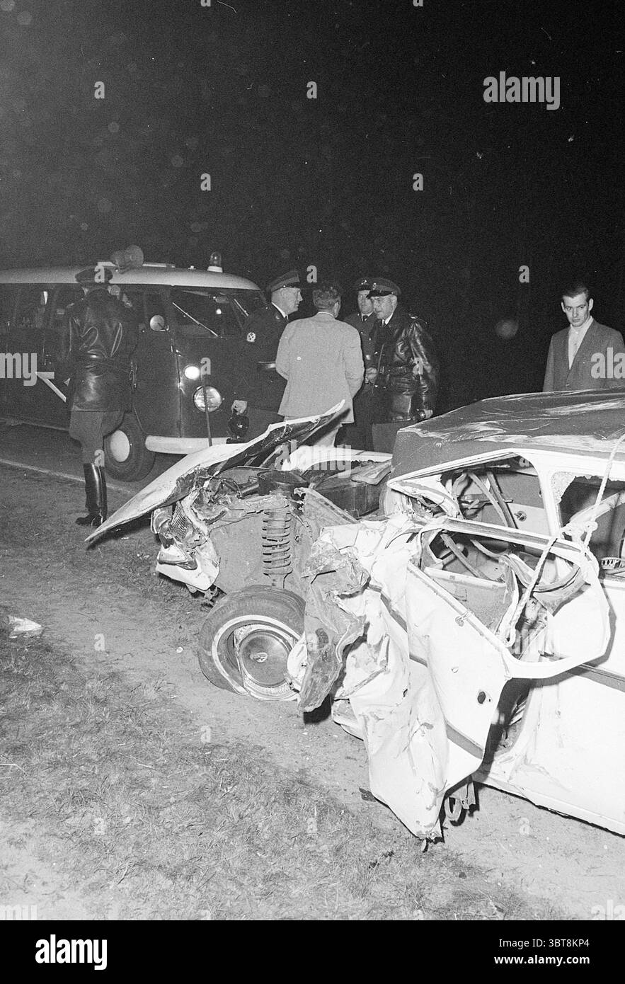 Incidente stradale relitti auto., Whizgle News, Dutch Desk, Paesi Bassi, 1950 - 2000. L'immagine mostra questi argomenti. La scena cattura un'impostazione notturna su una strada buia, illuminata solo dai fari di un veicolo nelle vicinanze. In primo piano, un'auto gravemente danneggiata si trova con la parte anteriore schiacciata e attorcigliata, rivelando metallo e detriti sparsi. Il colore dell'auto è per lo più oscurato dall'oscurità, ma alcuni accenni di luce suggeriscono che si tratta di una tonalità più leggera, forse bianca. Nelle vicinanze, un gruppo di individui, vestiti con un mix di abbigliamento formale e informale, sono riuniti lontano dal relitto Foto Stock