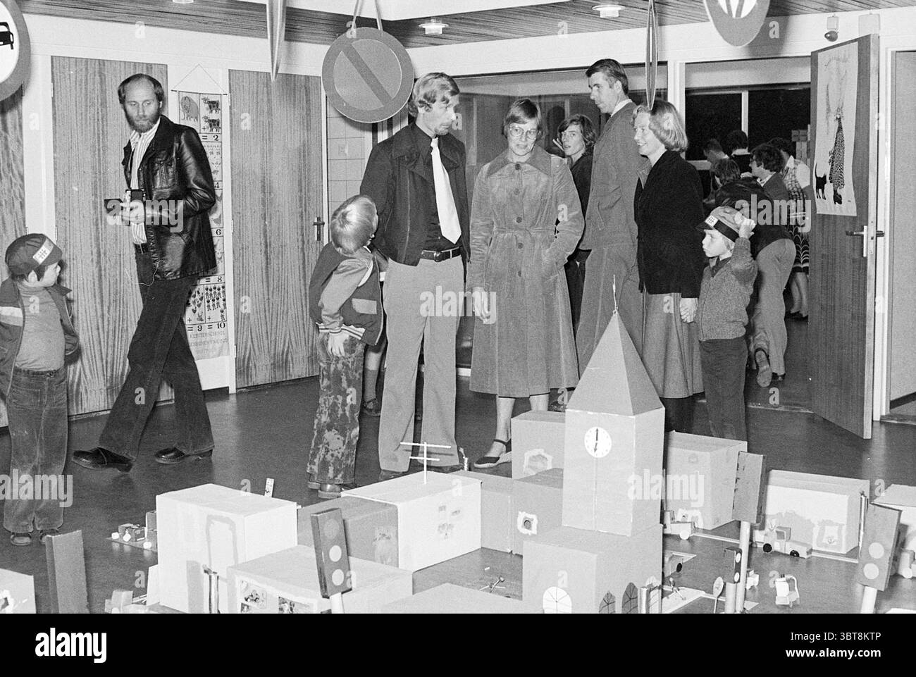 Traffic Exhibition Kindergarten Kennemer Joy Traffic Safe Traffic, Whizgle News, Dutch Desk, Paesi Bassi, 1950 - 2000 on 30-09-1976. L'immagine contiene questi argomenti. La scena cattura un ambiente interno vivace, probabilmente un centro comunitario o una scuola, pieno di attività. Il pavimento è liscio e scuro, fornendo un netto contrasto con le pareti più chiare e di colore neutro della stanza. Vari cartelli raffiguranti i simboli del traffico sono appesi al soffitto, aggiungendo un'atmosfera giocosa ma istruttiva. Un gruppo di adulti si riunisce al centro, impegnato nella conversazione, trasuda un senso di curiosità Foto Stock