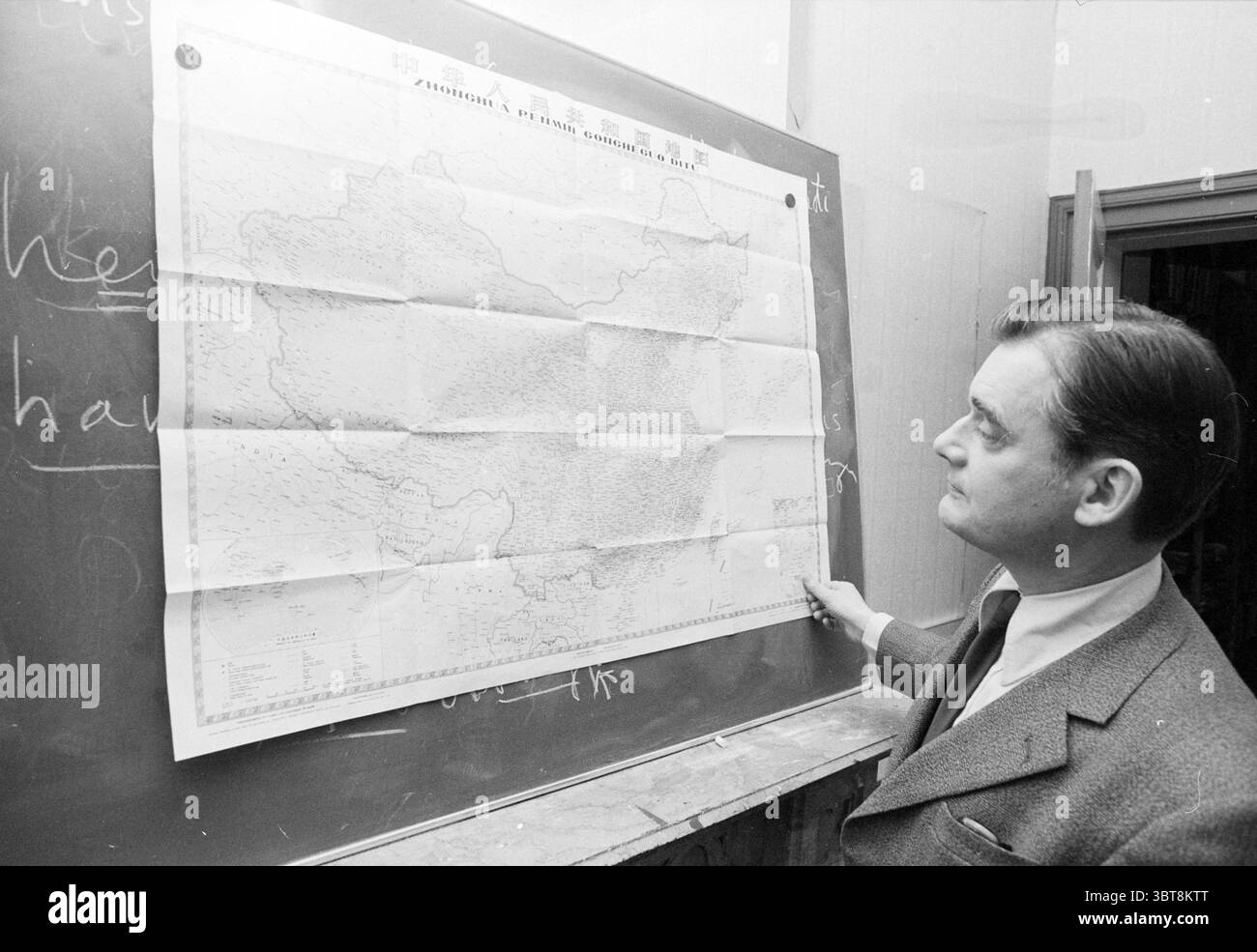 Uomo con carta cinese, Whizgle News, Dutch Desk, Paesi Bassi, 1950 - 2000 il 12-1978. Questi sono gli elementi dell'immagine. La scena vede un uomo in piedi accanto a una grande mappa aperta fissata a una lavagna scura. La mappa mostra caratteristiche geografiche dettagliate, tra cui montagne, fiumi e forse confini politici, resi in varie sfumature di grigio. L'ambiente sembra essere un'aula o un ufficio, indicato dalla presenza della lavagna, che presenta segni e contorni di gesso, che accennano a discussioni o lezioni precedenti. L'uomo è vestito con un abito sartoriale, comp Foto Stock
