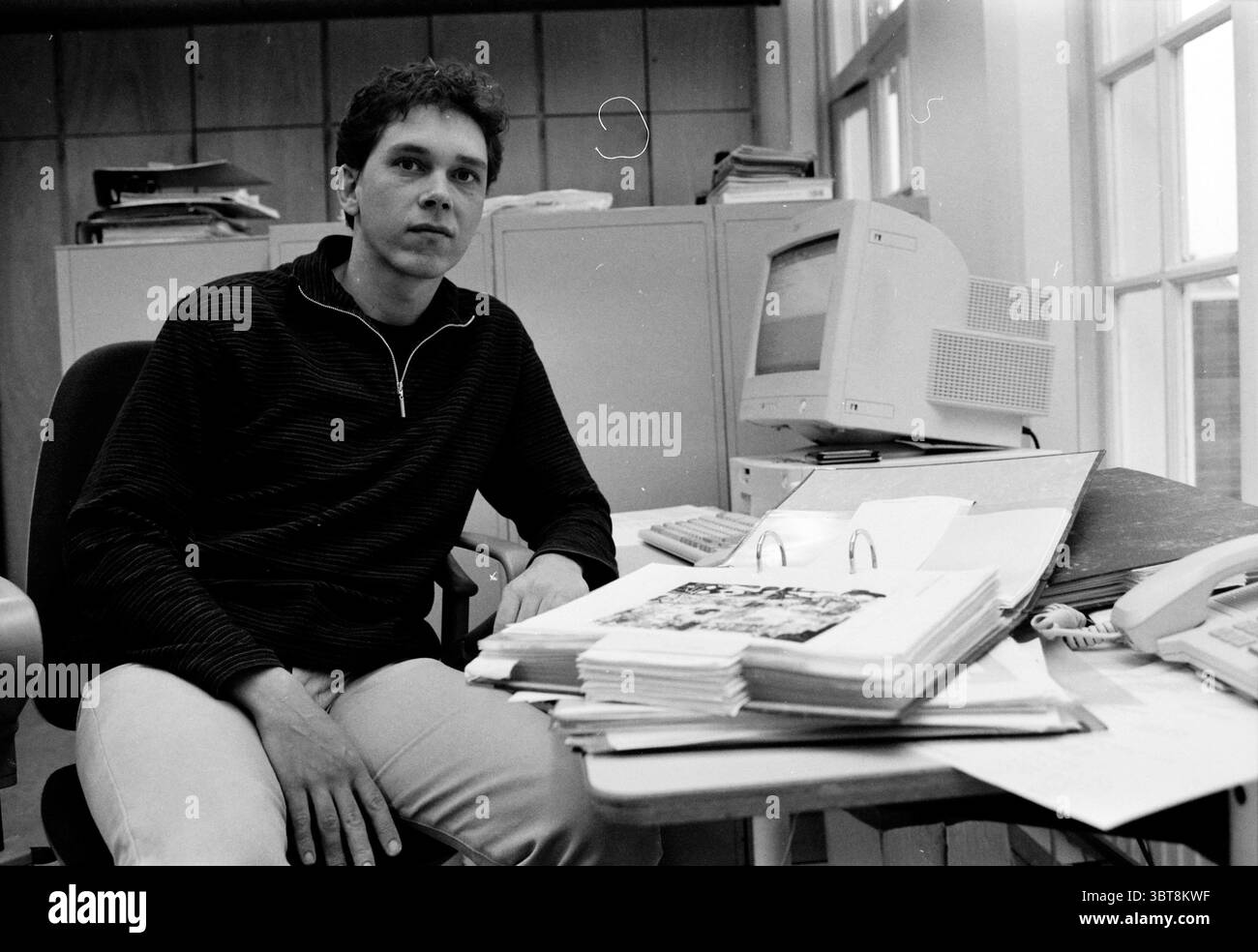 Frans Hals Museum Employees Haarlem Paesi Bassi, Whizgle News, Dutch Desk, Paesi Bassi, 1950 - 2000 il 24-11-1999. Questi argomenti sono mostrati nell'immagine. Nella scena, un giovane è seduto a una scrivania disordinata. Guarda direttamente lo spettatore con un'espressione composta. I suoi capelli sono scuri e leggermente stirati, e indossa un maglione nero su pantaloni di colore chiaro. La texture del maglione aggiunge profondità al suo aspetto e all'ambientazione più formale. La scrivania è piena di fogli di carta di varie dimensioni, alcuni impilati in modo casuale mentre altri sono aperti, rivelando il contenuto stampato. Un piccolo legame Foto Stock