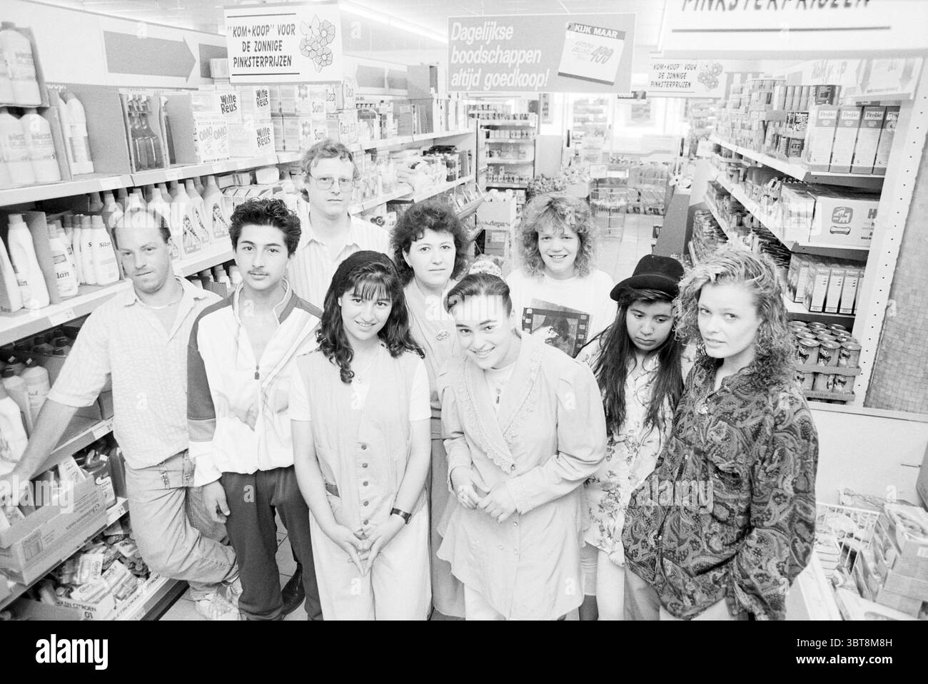 Dipendenti dell'azienda Kom + Koop, Whizgle News, Dutch Desk, Paesi Bassi, 1950 - 2000 il 26-05-1992. L'immagine include questi argomenti. In un corridoio ben rifornito, un gruppo diversificato di individui sta insieme, formando un accordo stretto. L'ambientazione è caratterizzata da ripiani luminosi pieni di una varietà di articoli per la casa e prodotti per la cura personale, creando uno sfondo vivace. Le persone nella scena sono un mix eclettico, che riflette stili ed espressioni diversi. Alcune persone indossano abiti casual, come camicie, camicette e abiti, con una miscela di colori che includono bianchi, blu Foto Stock