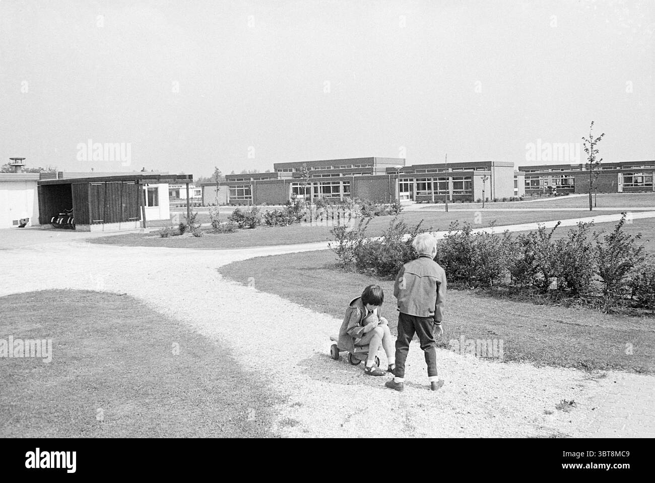 Scuola esterna Cruquiushoeve Exterior Schools, Whizgle News, Dutch Desk, Paesi Bassi, 1950 - 2000 il 22-05-1969. L'immagine mostra questi argomenti. La scena raffigura un ambiente tranquillo all'aperto, probabilmente intorno a una scuola o a una struttura educativa. In primo piano, due bambini sono il punto focale. Un bambino è seduto a terra, forse impegnato in un'attività come giocare o esaminare qualcosa da vicino, mentre l'altro sta accanto a loro, mostrando curiosità o interesse. Il paesaggio presenta un sentiero tortuoso fatto di ghiaia chiara che si snoda attraverso un'area erbosa verde, a indicare un pozzo-ma Foto Stock
