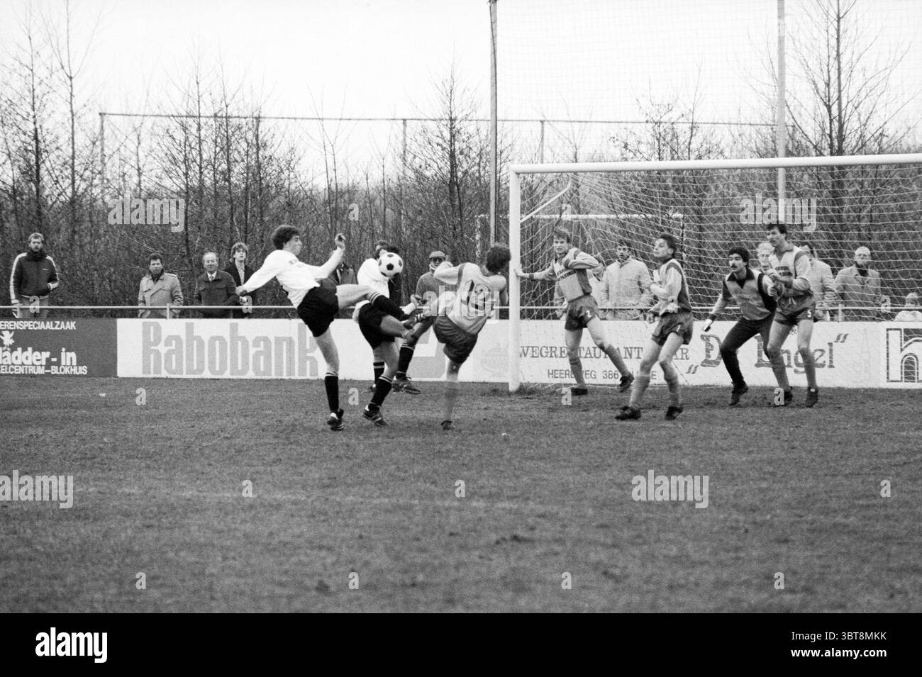 Terrasvogels - Hoofddorp Football, Whizgle News, Dutch Desk, Paesi Bassi, 1950 - 2000 il 21-12-1985. Questi sono gli argomenti nell'immagine. In una scena sportiva dinamica, due squadre sono impegnate in una partita di calcio vivace su un campo erboso, che mostra segni di usura e fango del gioco. I giocatori, vestiti con uniformi a contrasto, mostrano una gamma di colori con una squadra in una tonalità più scura e l'altra in una tonalità chiara. Il giocatore in primo piano, con indosso il nero, sta tentando abilmente di eseguire un calcio, la sua postura indica un momento di intensa concentrazione e sforzo. La palla, catturata mi Foto Stock
