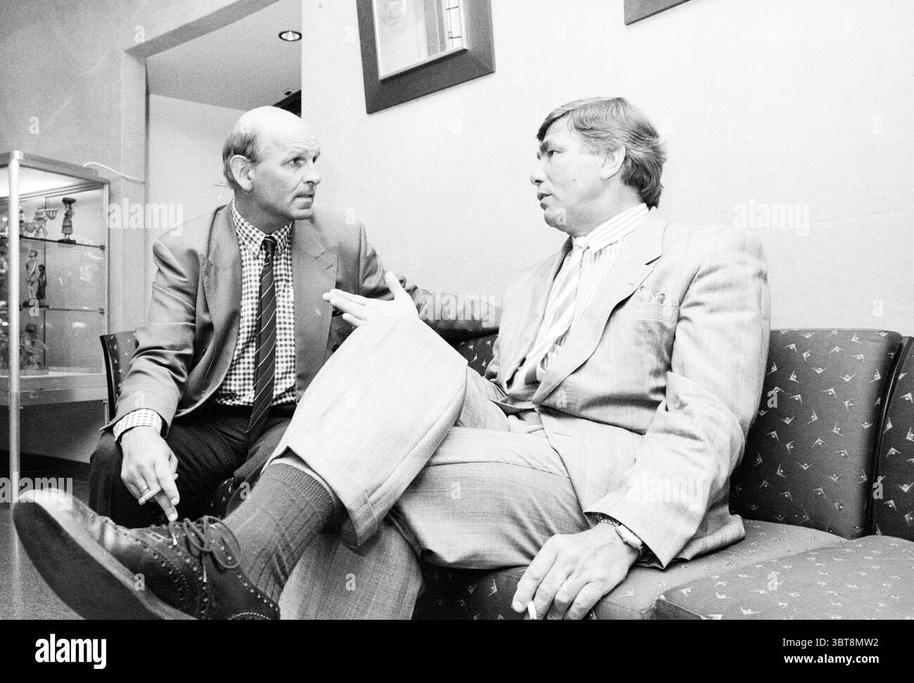 Due uomini, Whizgle News, Dutch Desk, Paesi Bassi, 1950 - 2000 il 05-1989. L'immagine include questi argomenti. In un ambiente silenzioso e monocromatico, due uomini sono impegnati in una conversazione mentre sono seduti su un divano a fantasia. La tavolozza dei colori si piega verso le sfumature di grigio, evocando un'atmosfera intramontabile e sofisticata. L'uomo sulla sinistra ha un taglio di capelli corti e indossa un abito sartoriale con una giacca di colore chiaro e una camicia a quadri, che mostra un'aria di formalità. Gesta leggermente con la mano destra, indicando il coinvolgimento nella discussione. La sua espressione è sincera, trasmette interesse e p Foto Stock
