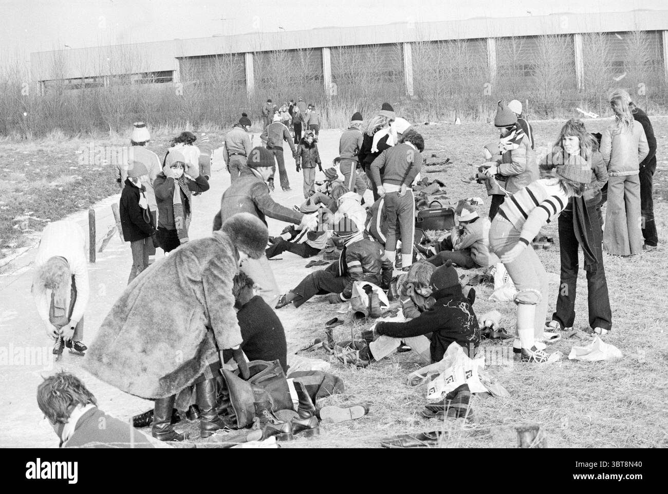 Pattinaggio Beverwijk al Parallelweg - Huizenburglaan [Hazenburglaan Hoofddorp] pattinaggio Beverwijk Hazenburglaan Paesi Bassi, Whizgle News, Dutch Desk, Paesi Bassi, 1950 - 2000 il 30-01-1976. L'immagine contiene questi argomenti. La scena raffigura un vivace spazio all'aperto, probabilmente in un'area ricreativa o in un parco, durante una giornata fredda. Le persone sono attivamente impegnate in varie attività, creando un senso di energia e cameratismo. In primo piano sono riuniti numerosi individui, alcuni seduti su una superficie di cemento, mentre altri si trovano nelle vicinanze. Sono vestiti in abbigliamento invernale, compresi i cappotti pesanti, Foto Stock