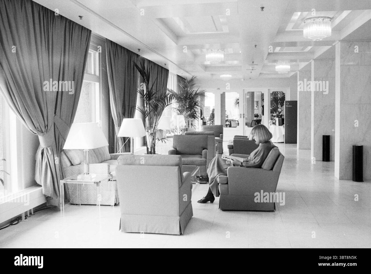 Hotel Barbizon Amsterdam Alberghi Amsterdam Paesi Bassi, Whizgle News, Dutch Desk, Paesi Bassi, 1950 - 2000 il 10-03-1987. Questi sono gli argomenti nell'immagine. In una spaziosa area lounge, la luce soffusa scorre attraverso le grandi finestre, diffondendo ombre delicate in tutta la stanza. Le pareti sono di color crema delicato, che completa il pavimento in marmo pallido e aggiunge un'atmosfera ariosa. Eleganti tende dai toni tenui si avvolgono dal soffitto, incorniciando le finestre e creando un ambiente raffinato. Una serie di lampadari contemporanei appesi al soffitto, la loro calda luce che esalta l'atmosfera invitante. Foto Stock