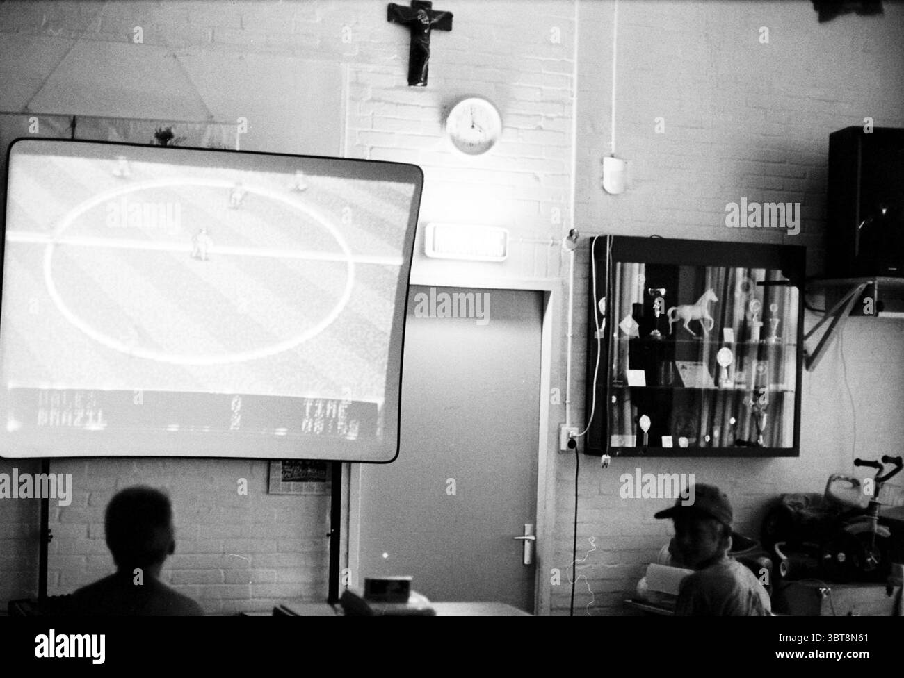 Nintendo final Haarlem Paesi Bassi, Whizgle News, Dutch Desk, Paesi Bassi, 1950 - 2000 il 04-08-1994. Questi argomenti sono mostrati nell'immagine. In una sala poco illuminata, un grande schermo domina lo spazio, emettendo un soffice bagliore che mette in risalto l'area circostante. Lo schermo visualizza un classico videogioco di calcio, con vivaci personaggi pixelati raggruppati su un campo verde, le loro posizioni suggeriscono un momento di gioco intenso. A sinistra, due persone si siedono sedute a disagio, mentre le figure si concentrano sullo schermo. L'illuminazione soffusa crea ombre profonde sulla loro f Foto Stock