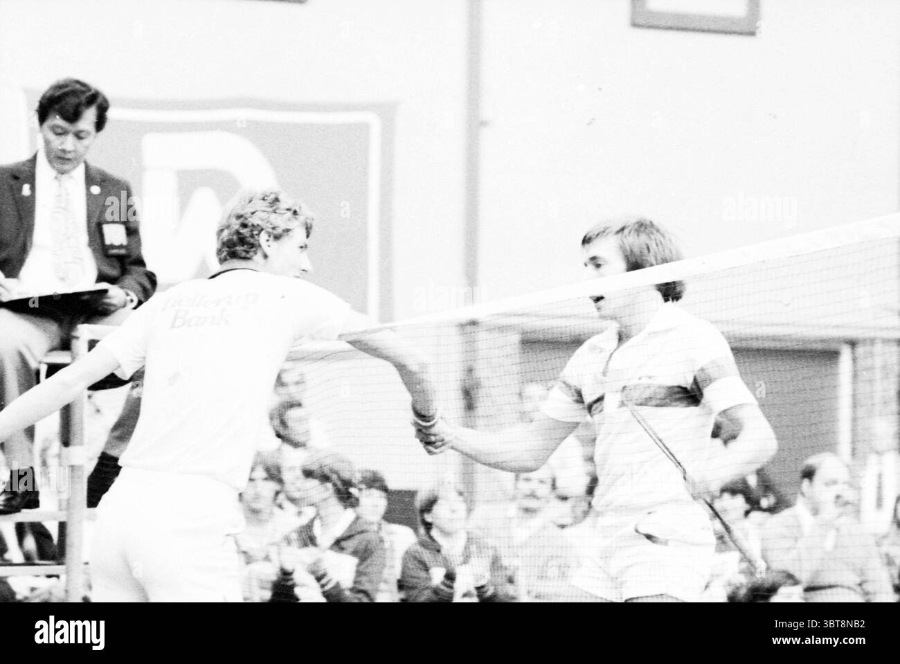 Partita di badminton maschile Duinwijckhal, Whizgle News, Dutch Desk, Paesi Bassi, 1950 - 2000 su 11-1979. Questi argomenti sono mostrati nell'immagine. La scena raffigura una vivace partita di tennis, che cattura un momento di sportività mentre due giocatori stringono la mano sulla rete. Un giocatore che indossa una camicia di colore chiaro presenta un colletto distinto e un'acconciatura riccia. L'altro giocatore indossa una camicia a righe con fasce orizzontali di varie tonalità pastello, evidenziando uno stile rilassato. Sullo sfondo, gli spettatori sono impegnati, con volti che appaiono attenti ed entusiasti. Il loro abbigliamento suggerisce un mix di cas Foto Stock