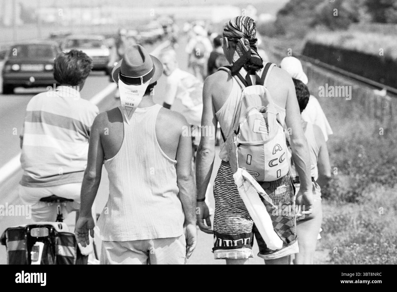 Miscellaneous Beach 6 - giorno, Whizgle News, Dutch Desk, Paesi Bassi, 1950 - 2000 il 28-07-1992. Questi argomenti sono mostrati nell'immagine. La scena si svolge lungo un percorso affollato accanto a una strada. Un mix di persone è visibile, camminando insieme in una processione informale. Due persone si distinguono in primo piano: Una indossa una camicia leggera senza maniche e un cappello a teglia larga, mentre l'altra sfoggia una bandana e porta con sé uno zaino. Le loro spalle sono rivolte verso lo spettatore, mostrando le loro posture rilassate. Il sentiero è delimitato da una vegetazione lussureggiante, che suggerisce il passaggio da urbano a naturale su Foto Stock