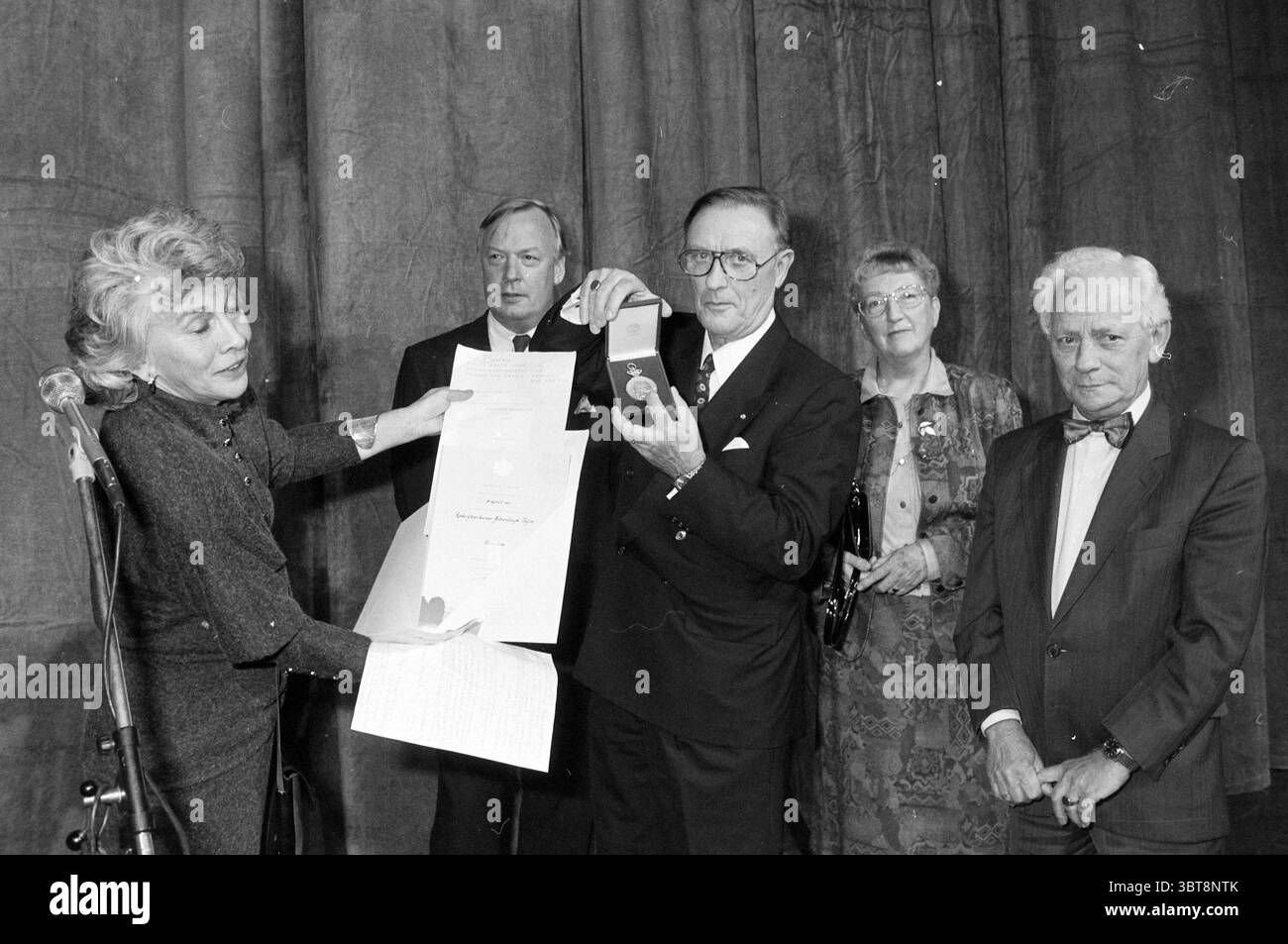 Medaglia onoraria Alberdingk Thijm associazione teatrale H'kerk Heemskerk, Whizgle News, Dutch Desk, Paesi Bassi, 1950 - 2000 il 10-11-1991. Questi argomenti vengono visualizzati nell'immagine. La scena cattura un evento formale, forse una cerimonia di premiazione, caratterizzato da un senso di celebrazione e riconoscimento. In primo piano, una donna si trova in posizione prominente a sinistra, con i suoi capelli a spalla elegantemente disegnati. Indossa un vestito oscuro e presenta un documento con una mano mentre tiene un medaglione premio nell'altra. La sua espressione trasmette serietà e orgoglio, indicando l'importanza del momento. Foto Stock