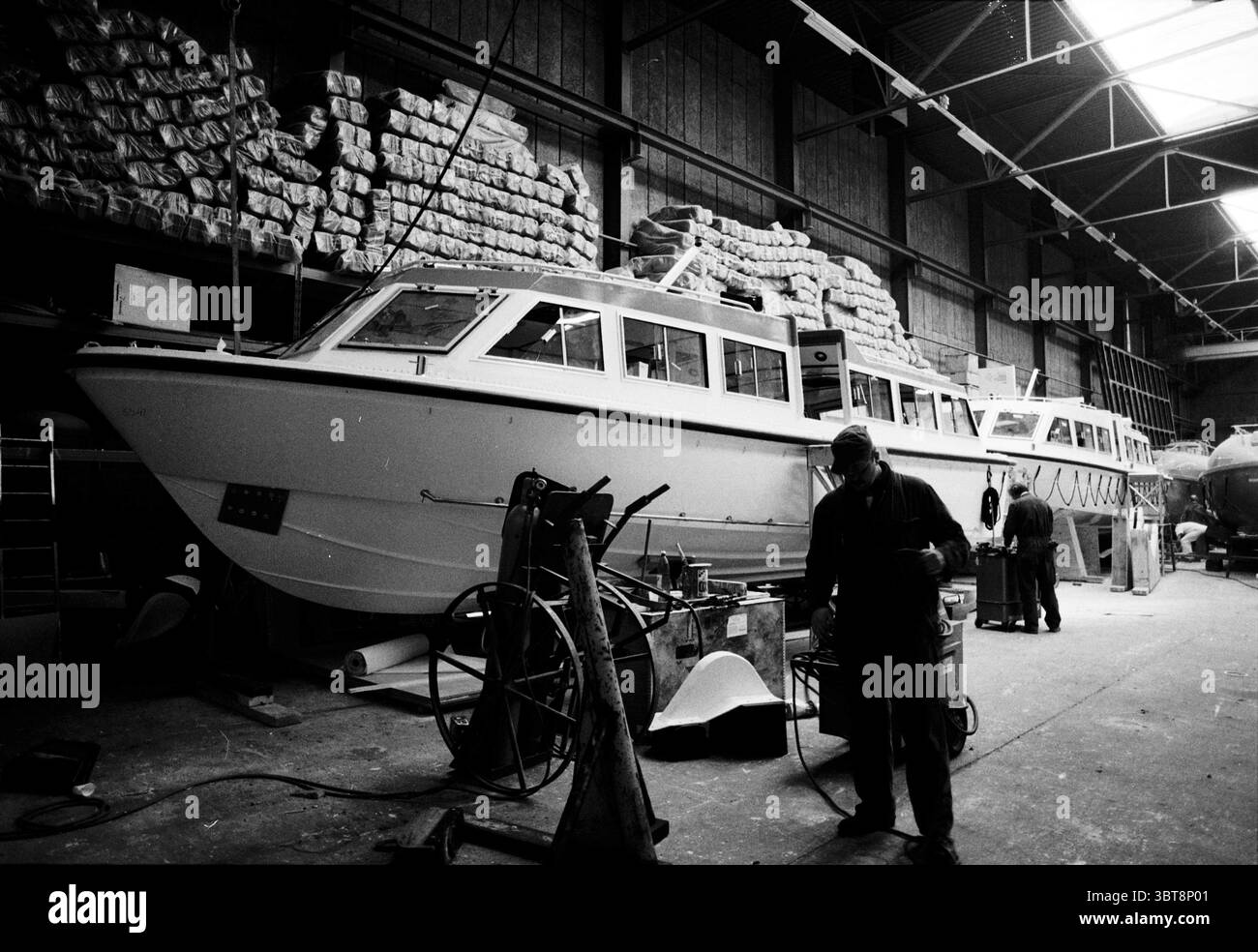 Costruzione di barche a motore a Mulder e Rijke IJmuiden IJmuiden Paesi Bassi, Whizgle News, Dutch Desk, Paesi Bassi, 1950 - 2000 il 03-11-1992. L'immagine mostra questi argomenti. In un ambiente industriale spazioso, una grande barca occupa una posizione prominente, il suo elegante scafo riflette la luce soffusa che filtra attraverso le alte finestre. L'esterno della barca è dipinto in un bianco frizzante, in contrasto con le tonalità più scure del magazzino circostante. Impilati sullo sfondo ci sono pile di materiali arrotolati, possibilmente teloni o tela, ordinatamente organizzati, che suggeriscono un livello di preparazione e o. Foto Stock