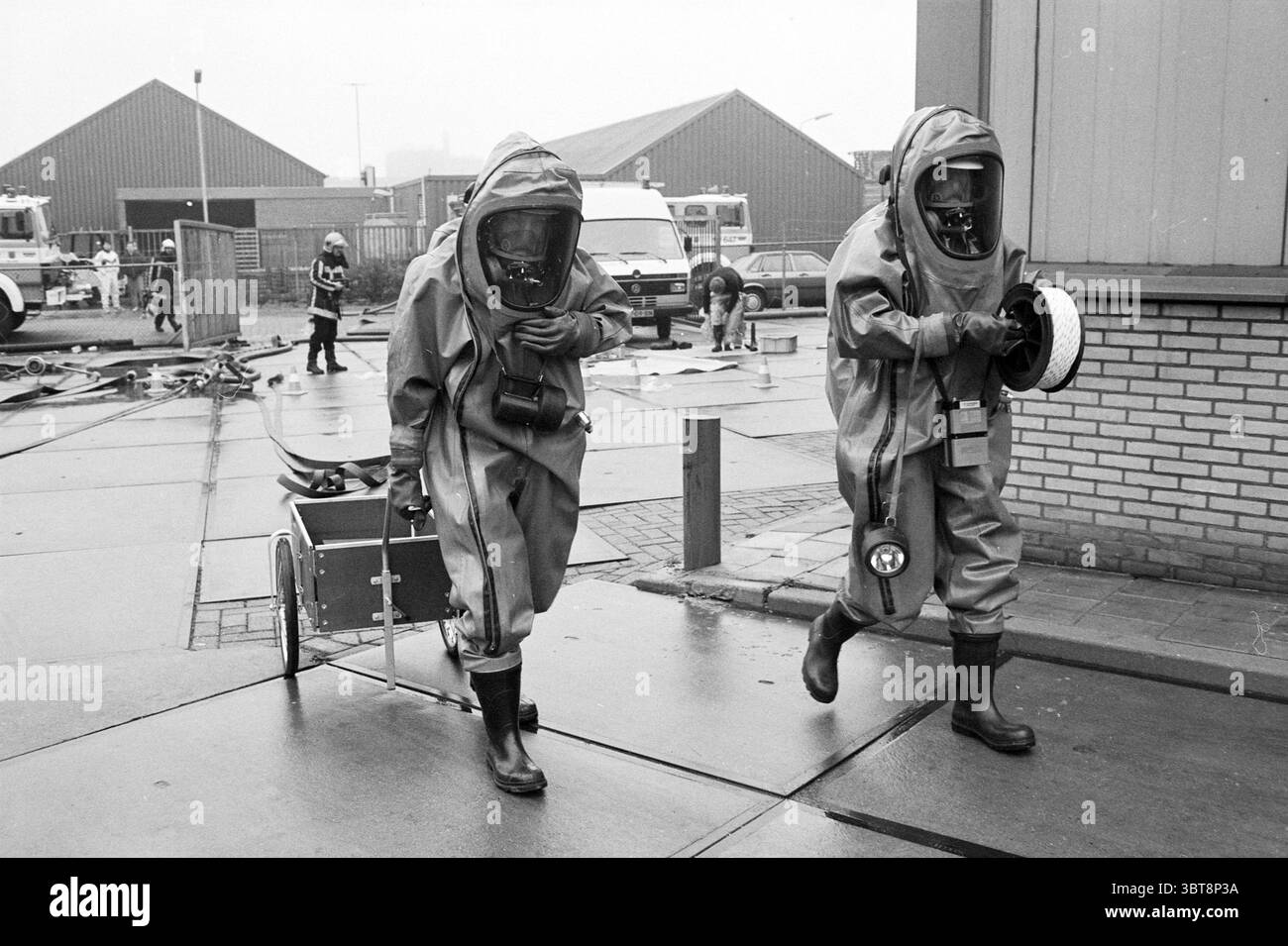 Esercitazione di controllo dell'ammoniaca Vigili del fuoco IJmuiden IJmuiden Paesi Bassi, Whizgle News, Dutch Desk, Paesi Bassi, 1950 - 2000 il 18-04-1993. Questi sono gli argomenti nell'immagine. In un ambiente grigio e industriale, due individui vestiti con tute protettive dominano la scena. Le loro tute, una sfumatura di giallo, sono progettate per proteggerle da materiali pericolosi. Ogni persona indossa un casco con visiera colorata che oscura il volto, aggiungendo un senso di anonimato e urgenza. Uno di essi tira un piccolo carrello, probabilmente dotato delle necessarie attrezzature di sicurezza, mentre l'altro porta un l Foto Stock