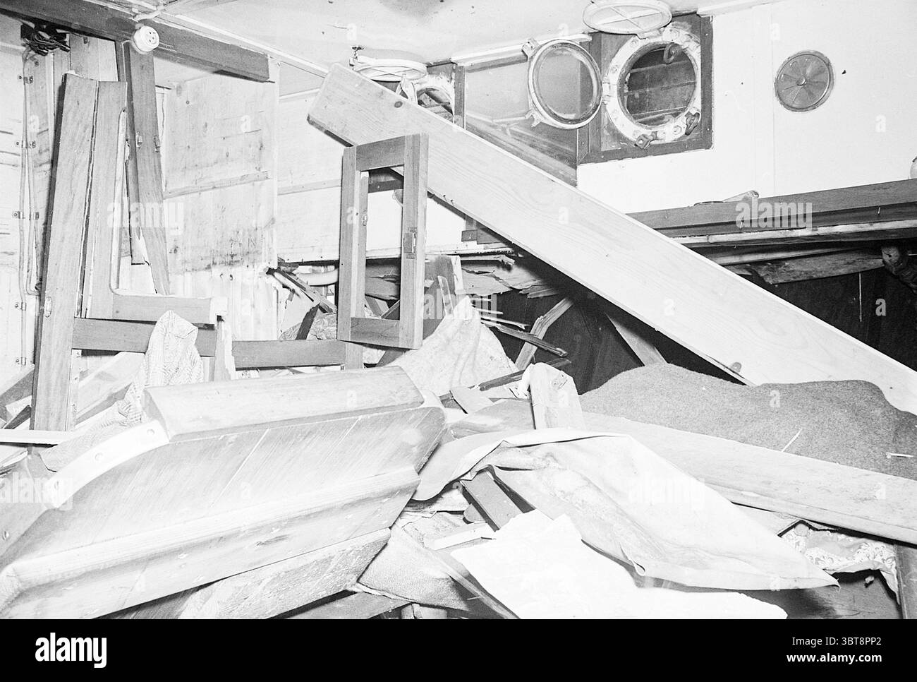 Katwijk 160 butano gas Explosion [a bordo della Vrouwe Alida] Explosions Ships, Whizgle News, Dutch Desk, Paesi Bassi, 1950 - 2000 il 06-04-1964. L'immagine mostra questi argomenti. La scena presenta un interno caotico, sopraffatto da un accumulo di detriti e disordine. Vari pezzi di legno, tra cui tavole e mobili, sono impilati e sparsi sul pavimento, creando un senso di confinamento e disordine. Il legno mostra una gamma di tonalità, dai marrone profondi alle tonalità più chiare, quasi sbiadite, suggerendo età e usura. Sullo sfondo, strutture deboli come muri e un par Foto Stock