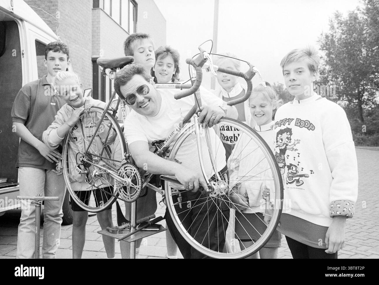 Squadra cronometro Mosquito round corriere team Joop van Ingen Mosquito round, Whizgle News, Dutch Desk, Paesi Bassi, 1950 - 2000 il 01-08-1987. L'immagine contiene questi argomenti. In una vivace scena all'aperto, un gruppo di adolescenti si riunisce intorno a un carismatico uomo che tiene una bicicletta. L'uomo, che indossa occhiali da sole sovradimensionati e un'espressione allegra, sembra dimostrare qualcosa di legato alla moto, catturando l'attenzione del pubblico giovane. La sua camicia è di colore chiaro, forse bianco, in contrasto con le sfumature più scure degli edifici circostanti. Il gruppo di sette adolescenti presenta una var Foto Stock
