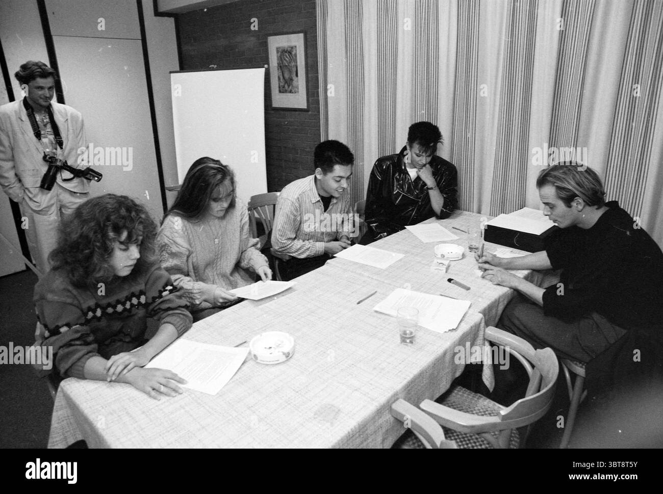 Agenzia di modelle Audition Hoofddorp Models Hoofddorp Paesi Bassi, Whizgle News, Dutch Desk, Paesi Bassi, 1950 - 2000 il 19-10-1989. Questi sono gli elementi dell'immagine. In un ambiente interno modesto, un gruppo di cinque persone è riunito intorno a un tavolo ricoperto da una tovaglia di colore chiaro. Lo sfondo è caratterizzato da una semplice parete dai toni neutri e da una tenda con un motivo morbido e verticale che consente di filtrare una luce delicata. Il primo individuo sulla sinistra ha capelli voluminosi e ricci e indossa un maglione decorato con un motivo colorato. Sembrano innestate, inclinate leggermente in avanti Foto Stock