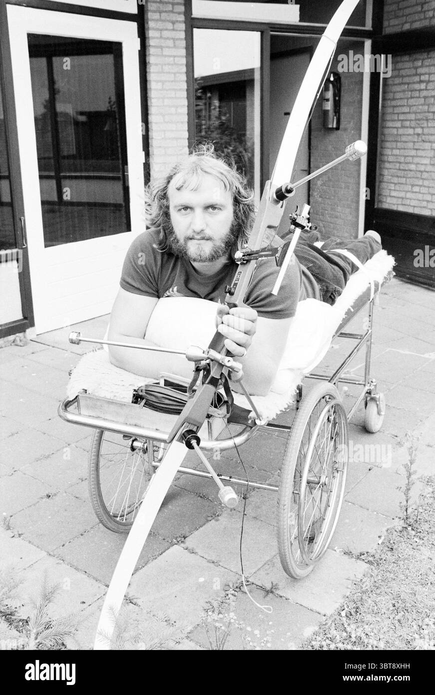 Lucas Schuring ha disabilitato archer Archery, Whizgle News, Dutch Desk, Paesi Bassi, 1950 - 2000 il 19-07-1978. Questi sono gli argomenti nell'immagine. In un'impressionante scena in bianco e nero, un uomo è in primo piano, mentre si rilassa sulla cima di un dispositivo a ruote simile a una sedia a rotelle improvvisata. La sua postura è rilassata ma decisa quando si afferra un fiocco, le sue linee eleganti contrastano con il design più pratico del carrello. I lunghi capelli dell'uomo cadono intorno al viso, aggiungendo un carattere robusto alla composizione. Lo sfondo offre uno scorcio architettonico di un edificio con grandi finestre rettangolari, r Foto Stock