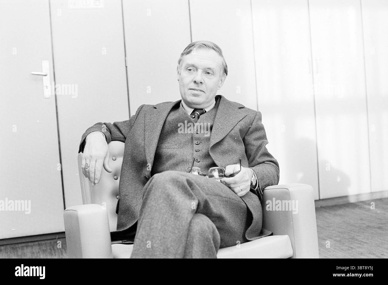 Intervista il sindaco Reehorst Velsen Mayors, Whizgle News, Dutch Desk, Paesi Bassi, 1950 - 2000 il 23-12-1976. L'immagine contiene questi argomenti. In un interno con luce soffusa, un uomo siede comodamente su una sedia imbottita, che presenta bordi arrotondati e un colore chiaro e neutro. È vestito con una tuta formale in tre pezzi, prevalentemente in tonalità scura, accentuata da un motivo sottile che aggiunge texture senza compromettere l'aspetto generale. Il suo comportamento è calmo e composto, trasmettendo un'aria di meditazione. Tiene un piccolo oggetto metallico, forse una tazza o un bicchiere, in una mano, suggerendo di essere Foto Stock