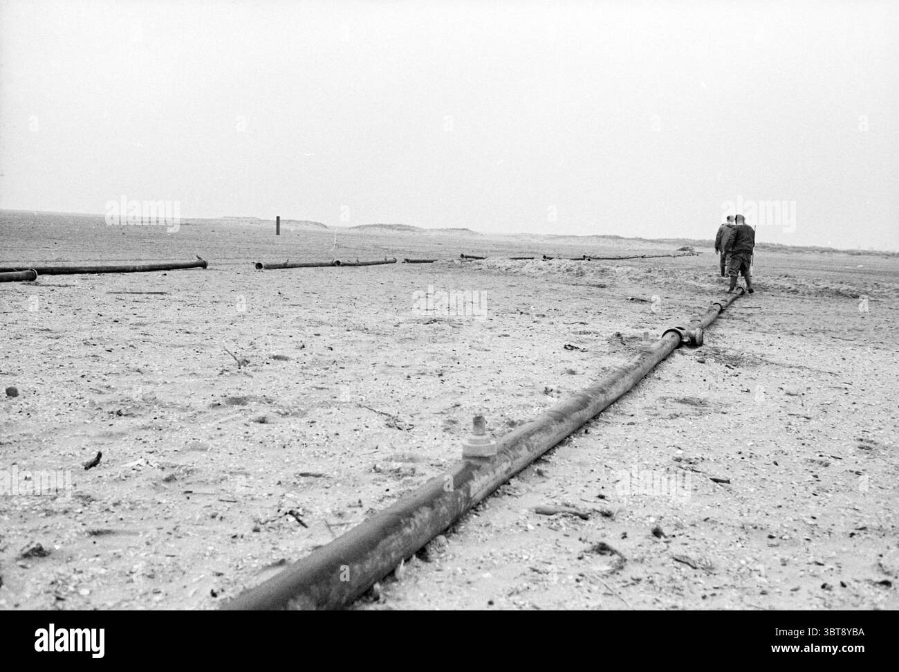 Costruzione di oleodotti IJmuiden Beach Pielines Spiaggia e spiagge IJmuiden Paesi Bassi, Whizgle News, Dutch Desk, Paesi Bassi, 1950 - 2000 il 16-02-1982. Questi sono gli elementi dell'immagine. In un paesaggio arido, si vedono due figure camminare lungo una lunga struttura tubolare che serpeggia attraverso il terreno sabbioso. Il terreno è prevalentemente un beige tenue, con macchie di sabbia grossolana e piccole pietre sparse intorno, contribuendo a creare un'atmosfera quasi desolata. Il cielo sovrastante è coperto, emettendo una luce grigia opaca che ammorbidisce le ombre e crea un'atmosfera malinconica. Le cifre, dres Foto Stock