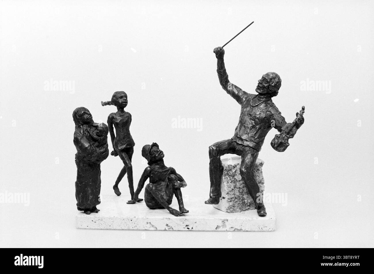 Piccolo gruppo di sculture, Whizgle News, Dutch Desk, Paesi Bassi, 1950 - 2000 il 10-1984. Questi sono gli argomenti nell'immagine. La scena presenta una scultura in bronzo che raffigura un momento coinvolgente tra un conduttore e tre figure. Il conduttore, seduto su un blocco, tiene la bacchetta di un direttore in una mano e un violino nell'altra. La sua postura è animata, trasmettendo un senso di passione ed energia, come se stesse guidando un'orchestra invisibile. Alla sua sinistra, una donna culla un bambino vicino a lei, il suo sguardo diretto verso il direttore d'orchestra, suggerendo ammirazione o gioia. Indossa indumenti fluenti Foto Stock