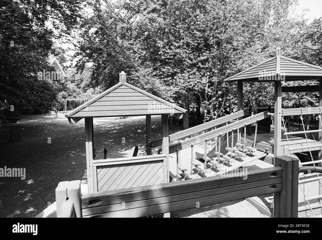 Playground Forest and Dune School Bloemendaal Bos en Duinlaan, Whizgle News, Dutch Desk, Paesi Bassi, 1950 - 2000 il 25-06-1996. L'immagine contiene questi argomenti. La scena è ambientata in un parco giochi circondato da una vegetazione lussureggiante, creando un'atmosfera vibrante e invitante. L'area è per lo più ombreggiata da alberi alti, il loro fitto fogliame che filtra la luce del sole per creare macchie di luce e ombra sul terreno. In primo piano, strutture in legno formano un'area giochi, caratterizzata da due piccoli padiglioni o gazebo con tetti leggermente inclinati e ringhiere in legno. Il legno è di una tonalità calda, anche se Foto Stock
