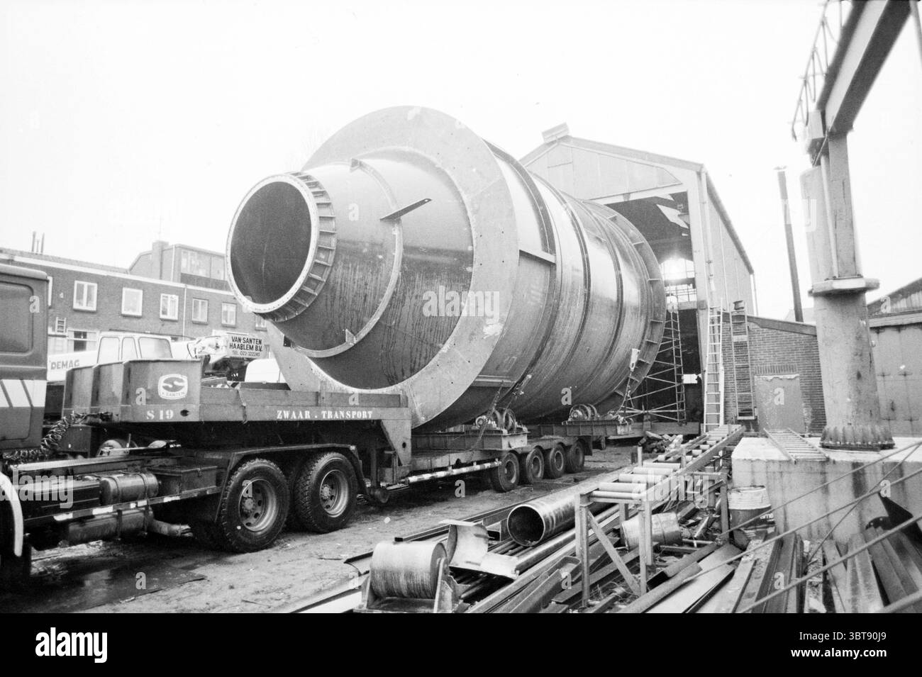 Grande caldaia sul camion. Aziende di trasporto, Whizgle News, Dutch Desk, Paesi Bassi, 1950 - 2000 il 02-03-1987. Questi sono gli elementi dell'immagine. La scena presenta una grande configurazione industriale dominata da un massiccio oggetto cilindrico, presumibilmente un componente di un macchinario o parte di un processo di produzione. Il cilindro, metallico e leggermente riflettente, è posizionato su un carro attrezzi con pianale che suggerisce che è in transito o è appena arrivato. L'ambiente circostante è composto da un ambiente di fabbrica o magazzino, caratterizzato da strutture precedentemente menzionate parzialmente visibili nel Foto Stock