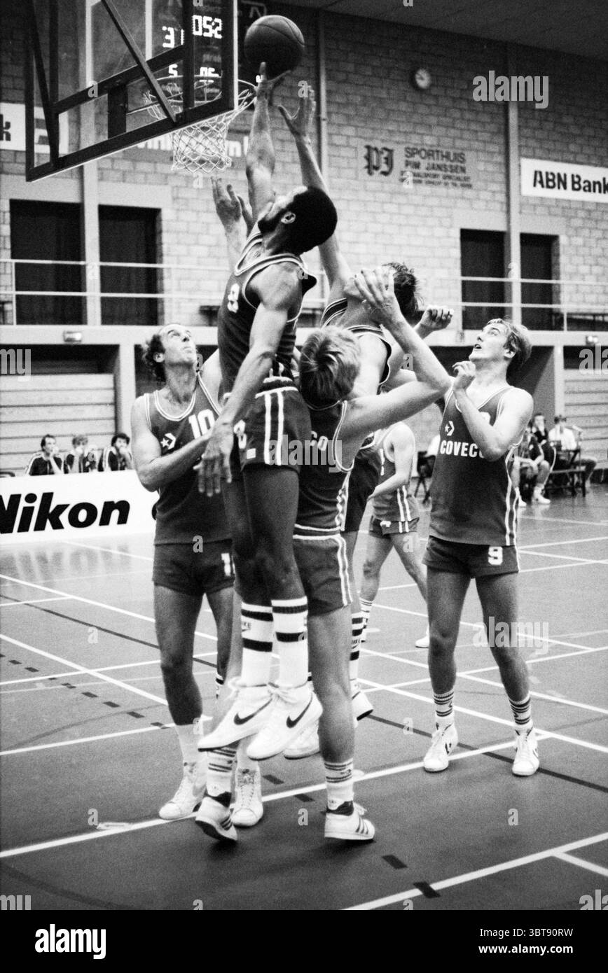 Partita di basket., Whizgle News, Dutch Desk, Paesi Bassi, 1950 - 2000. Questi sono gli elementi dell'immagine. La scena cattura un momento di basket dinamico in una palestra piena di attività. Al centro, un giocatore salta in alto verso il cerchio, il braccio si estende mentre si prepara a fare un tiro. La sua uniforme, con strisce audaci e il numero 9, contrasta vividamente con l'abbigliamento più scuro dei difensori che lo circondano. I giocatori che lo circondano mostrano espressioni intense, sottolineando la natura competitiva del gioco. Un giocatore, che indossa il numero 10, sta tentando di bloccare il tiro mentre altri, Foto Stock