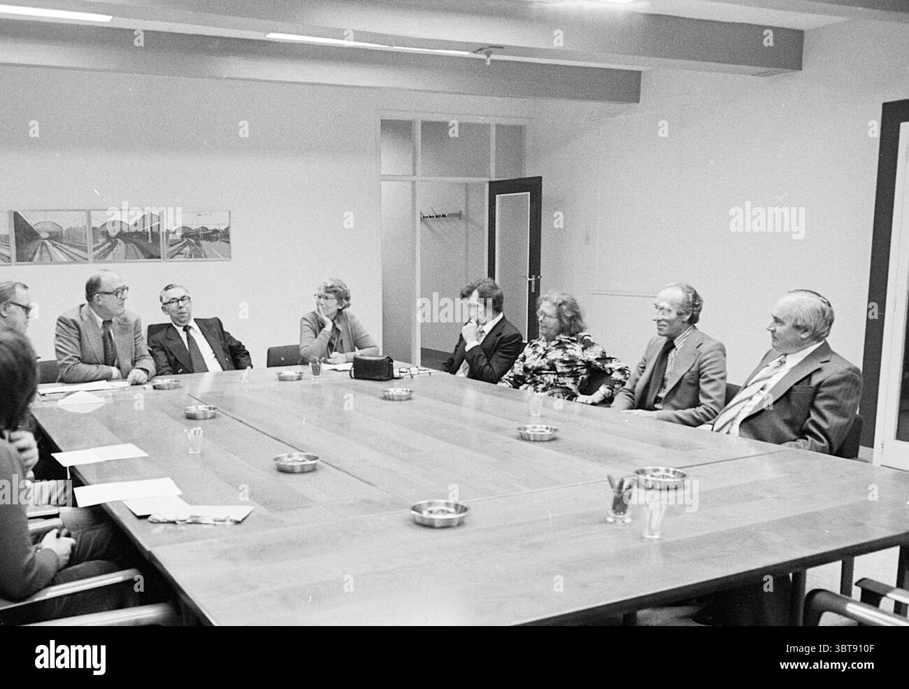 Installazione Meeting Temporary Housing Foundation Housing Installation Meetings, Whizgle News, Dutch Desk, Paesi Bassi, 1950 - 2000 il 14-09-1976. L'immagine mostra questi argomenti. La scena raffigura un incontro formale che si svolge intorno a un grande tavolo da conferenza in legno, che ha una superficie lucida ed è allestito in una sala altrimenti moderna e decorata minimamente. Il tavolo è rettangolare con una finitura a grana naturale, e sulla sua superficie sono presenti diversi posacenere, a indicare che il fumo potrebbe aver avuto luogo. Intorno al tavolo, sono seduti otto persone, ciascuna impegnata in una conversazione Foto Stock