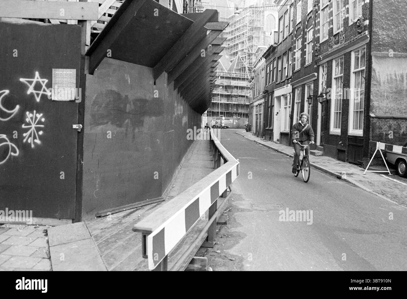 Ponte pedonale in legno Jansstraat Hout Straten Haarlem Jansstraat Paesi Bassi, Whizgle News, Dutch Desk, Paesi Bassi, 1950 - 2000 il 01-12-1983. Questi sono gli argomenti nell'immagine. La scena raffigura una strada stretta e tortuosa racchiusa da edifici su entrambi i lati. L'architettura mostra un mix di fascino del vecchio mondo con facciate in mattoni ed elementi di costruzione moderni come impalcature sullo sfondo. Sul lato sinistro, c'è una struttura prominente con un cordolo allungato che si curva drammaticamente, creando un senso di recinzione. Questa funzione ha una trama che suggerisce che si tratta di un'installazione temporanea Foto Stock