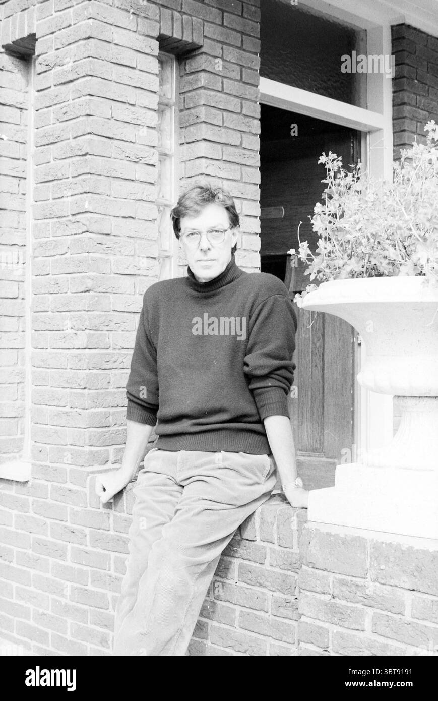 Frans Molenaar in Zandvoort Zandvoort, Whizgle News, Dutch Desk, Paesi Bassi, 1950 - 2000 il 24-09-1990. L'immagine mostra questi argomenti. In un ambiente caratterizzato da muri in mattoni rossi testurizzati, una figura è casualmente appollaiata su una sporgenza bassa. La persona che indossa un maglione dolcevita scuro che contrasta con i toni più chiari dell'ambiente circostante, si appoggia leggermente con una postura rilassata. I loro capelli appaiono arruolati e sono incorniciati dall'ingresso dietro di loro, il che suggerisce un interno accogliente. Intorno a loro, è visibile un grande vaso di fiori, che ospita una vegetazione lussureggiante che rompe l'ateo monocromatico Foto Stock