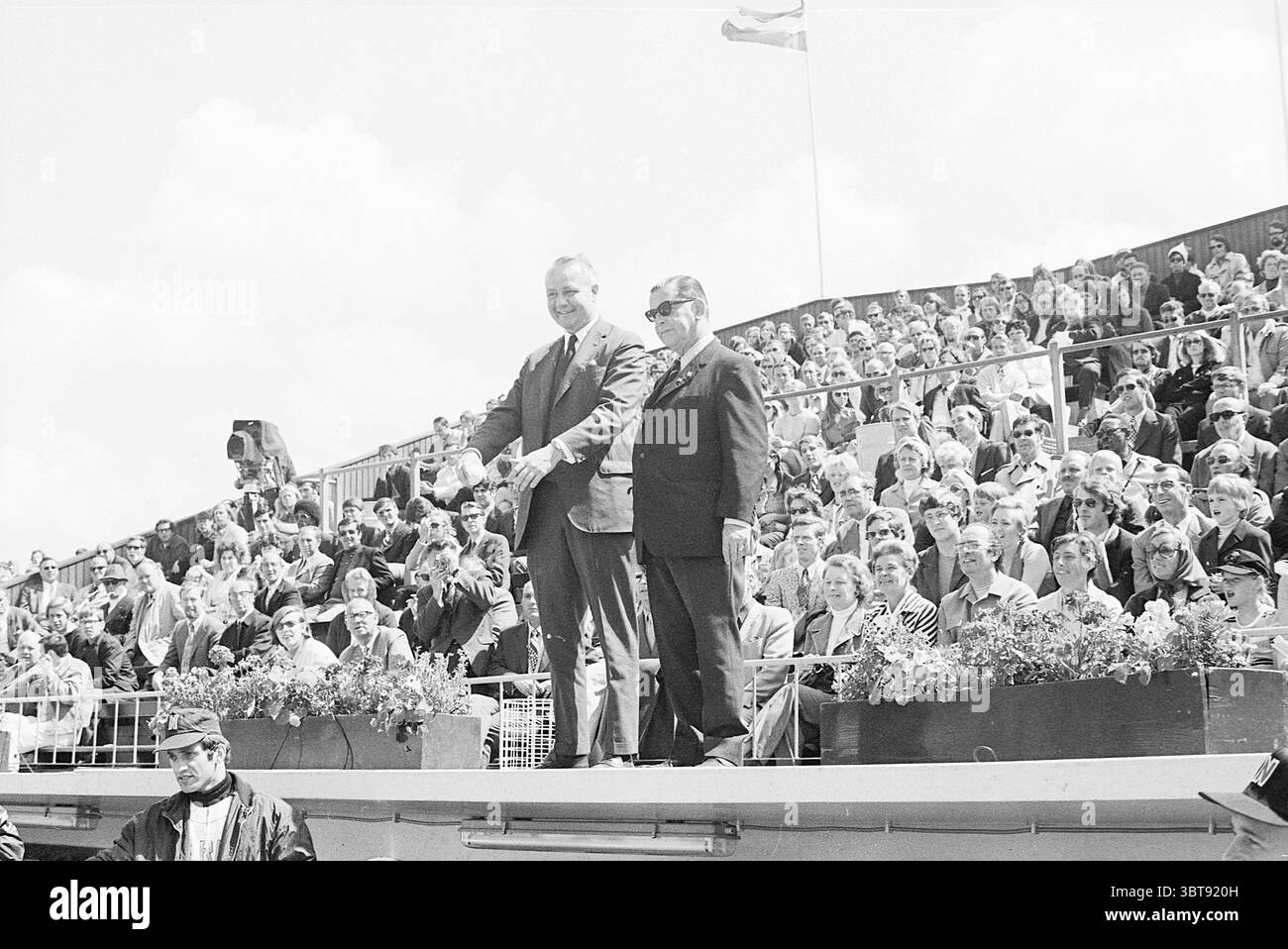 Baseball Paesi Bassi - Detroit ITM, Whizgle News, Dutch Desk, Paesi Bassi, 1950 - 2000 il 27-06-1971. Questi argomenti sono mostrati nell'immagine. In un vivace ambiente all'aperto, due uomini stanno in piedi in modo prominente su una piattaforma rialzata, le loro espressioni sono un mix di orgoglio e gioia. Entrambi sono vestiti con un abbigliamento formale, con abiti aderenti che trasmettono un senso d'occasione. Un uomo ha alzato la mano come se riconoscesse la folla, mentre l'altro sta al suo fianco, impegnato in un momento di cameratismo. Lo sfondo è pieno di un vasto pubblico, seduto a livelli, che rivela un'assemblea diversificata di individui. Il cro Foto Stock