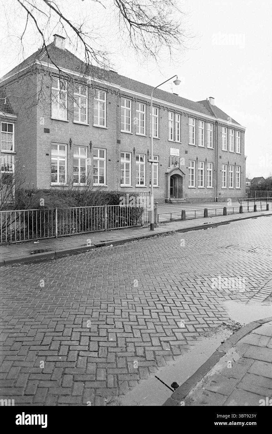 Esterno Cassianusschool B'wijk Exterior Schools Beverwijk Romerkerkweg Paesi Bassi, Whizgle News, Dutch Desk, Paesi Bassi, 1950 - 2000 il 03-12-1976. L'immagine contiene questi argomenti. La scena cattura un edificio scolastico in mattoni che si estende su più piani, caratterizzato da grandi finestre divise da griglie, consentendo l'ingresso di molta luce naturale. L'architettura presenta un design simmetrico con tetti a spiovente e un'entrata prominente, incorniciata da una porta ad arco. Di fronte all'edificio, un pavimento bagnato risplende, riflettendo le sfumature di grigio del cielo coperto. Il ciottolo Foto Stock