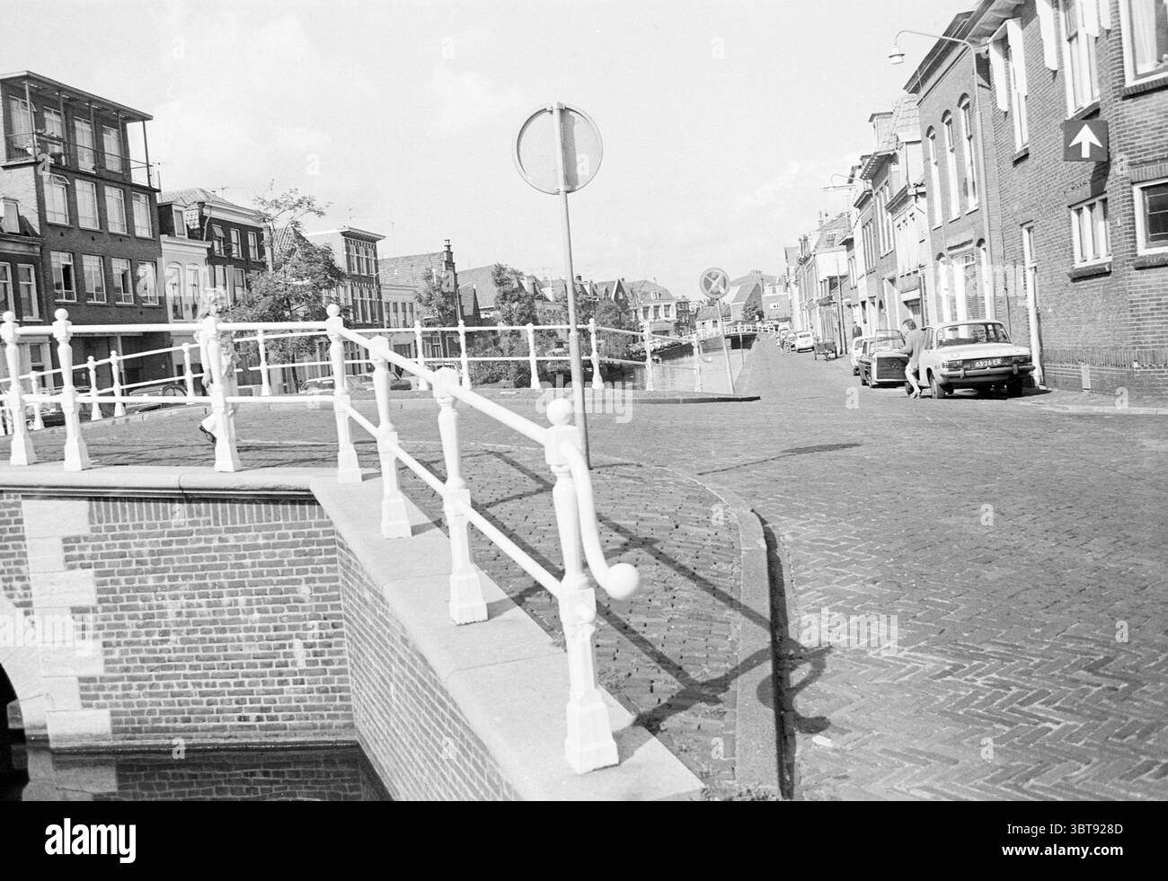 Vieni a visitare il Burgwal Haarlem Paesi Bassi, Whizgle News, Dutch Desk, Paesi Bassi, 1950 - 2000 al numero 25-08-1975. L'immagine include questi argomenti. La scena raffigura un tranquillo ambiente urbano caratterizzato da una strada dolcemente tortuosa fiancheggiata da edifici in mattoni. Sulla sinistra, un canale affascinante risplende delicatamente, riflettendo la luce soffusa. Una ringhiera bianca curva elegantemente lungo il bordo del canale, migliorando la pittoresca qualità dell'ambiente. Gli edifici presentano un mix di architettura tradizionale e moderna, caratterizzate da toni terrosi di marrone e grigi tenui che si fondono armoniosamente con il Foto Stock