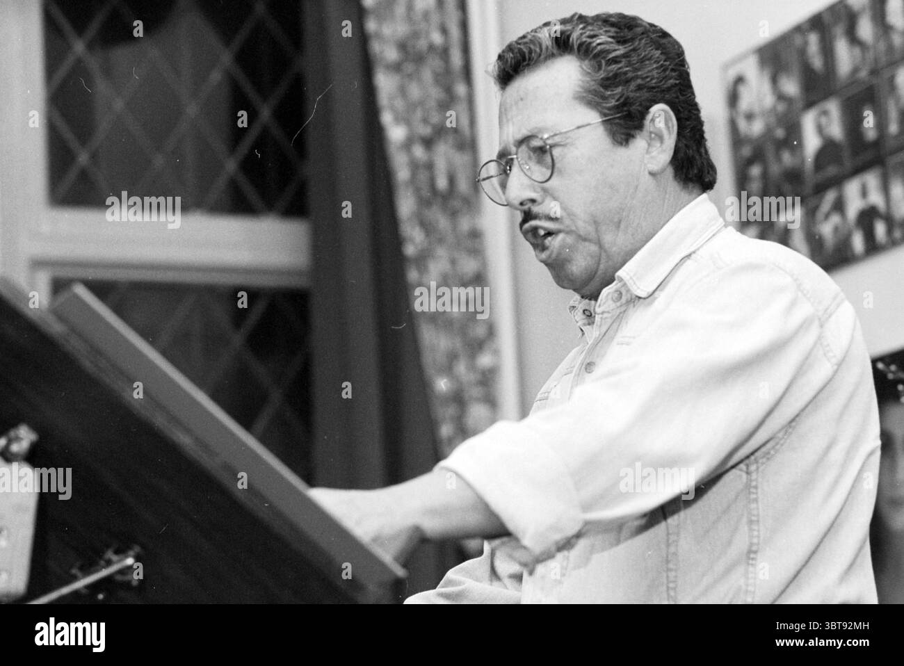 Casa nostra Singing Evening, Whizgle News, Dutch Desk, Paesi Bassi, 1950 - 2000 on 27-09-1996. Questi sono gli argomenti nell'immagine. Un uomo è seduto in un ambiente interno accogliente, sembra impegnarsi appassionatamente nell'atto di esibirsi o presentare. È vestito con una camicia e un paio di occhiali di colore chiaro, la sua espressione è focalizzata e animata. I suoi capelli sono leggermente ruvidi e le sue caratteristiche facciali mostrano una miscela di concentrazione ed entusiasmo. Lo sfondo è decorato con ricche texture, tra cui tende a motivi geometrici che incorniciano una finestra con un design a reticolo diamantato, che consente di filtrare una luce soffusa Foto Stock