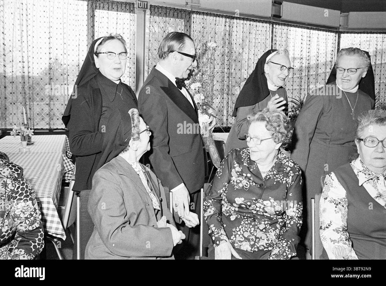 Lode Bob Scholten Treslong Hillegom loda persone che cantano e musica, Whizgle News, Dutch Desk, Paesi Bassi, 1950 - 2000 il 24-02-1977. Questi sono gli argomenti nell'immagine. La scena cattura un incontro in uno spazio interno ben illuminato, probabilmente un banchetto o una sala comune, caratterizzato dalla sua calda atmosfera e da un arredamento discreto. In primo piano, un gruppo di donne anziane siedono vicino a un tavolo adornato con una tovaglia a scacchi, creando un'atmosfera invitante e comune. Il loro abbigliamento è vario, con uno che indossa una camicetta colorata e occhiali floreali, mentre altri sfoggiano motivi classici e neut Foto Stock