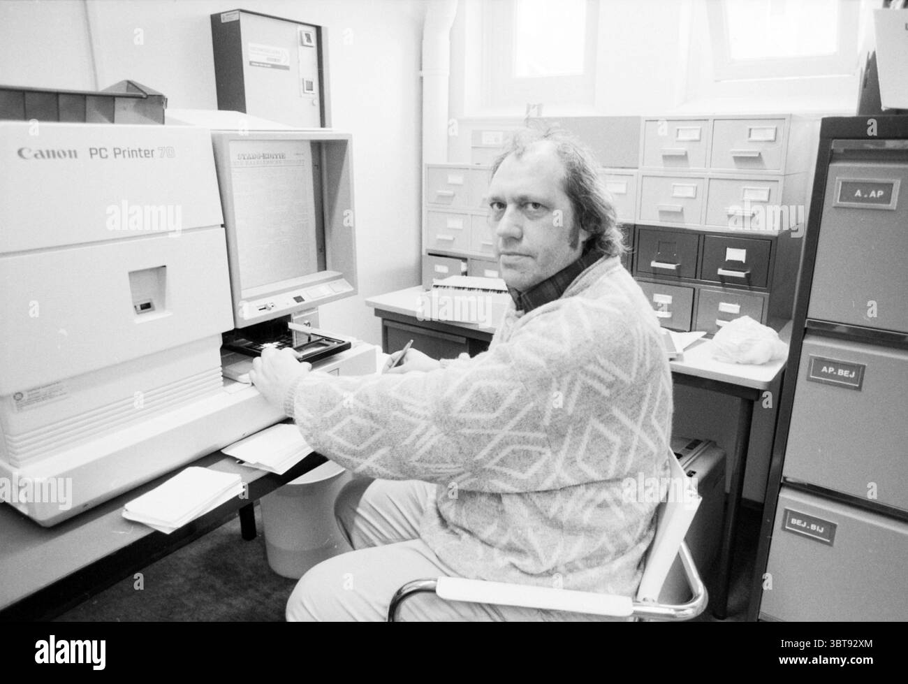 Vermooten effettua ricerche sulle ferrovie di Haarlem Railways, Whizgle News, Dutch Desk, Paesi Bassi, 1950 - 2000 il 21-01-1989. Questi argomenti vengono visualizzati nell'immagine. La scena raffigura un uomo seduto a una scrivania in un ufficio disordinato. Ha i capelli di media lunghezza e indossa un maglione a motivi leggeri che contrasta con l'aspetto utilitaristico dell'ambiente circostante. La sua espressione è seria, riflette la concentrazione o forse l'introspezione mentre guarda direttamente allo spettatore. La scrivania è occupata da una stampante Canon, che si distingue in modo prominente per la sua voluminosità e il colore bianco distinto, suggerisci Foto Stock