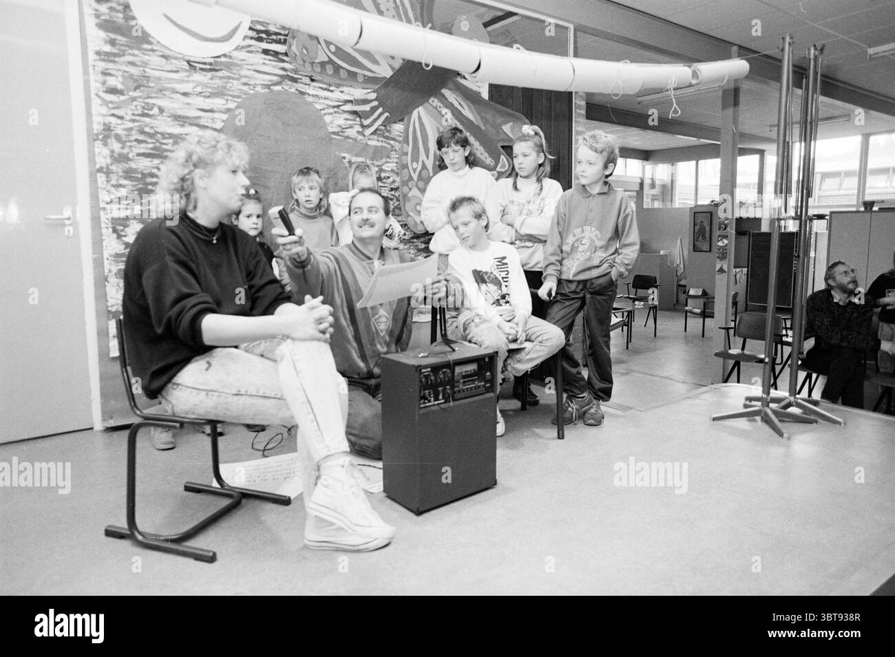 Overh. Money Pleiadenschool IJmuiden Handover and Transfers, Whizgle News, Dutch Desk, Paesi Bassi, 1950 - 2000 il 10-11-1988. Questi sono gli argomenti nell'immagine. In una sala luminosa e spaziosa piena di luce naturale, un gruppo di bambini siede con attenzione, riuniti intorno a una figura adulta che sembra coinvolgerli. L'adulto, con baffi e abbigliamento casual, tiene in mano un foglio di carta e gesti animati, attirando l'attenzione dei bambini. È seduto su una piccola piattaforma o sgabello, il che gli dà una posizione leggermente elevata rispetto al resto del gruppo. I bambini, un mix di ragazzi Foto Stock