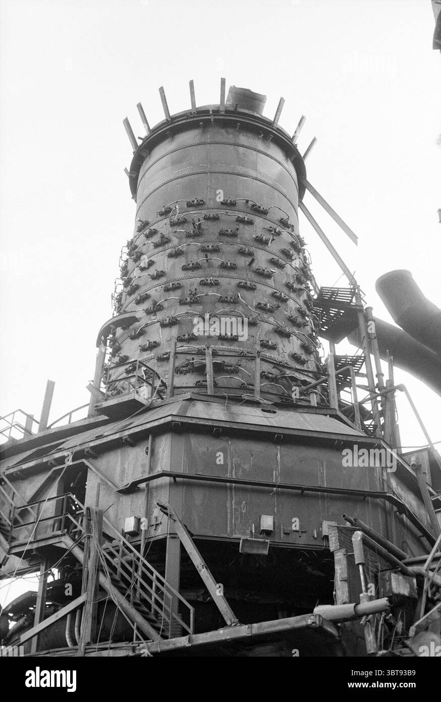 Demolizione/demolizione altoforno 3, Whizgle News, Dutch Desk, Paesi Bassi, 1950 - 2000 il 17-10-1991. L'immagine mostra questi argomenti. La scena presenta un'impressionante struttura industriale, torreggiante in primo piano contro un cielo pallido. La caratteristica principale è una grande sezione cilindrica sormontata da sporgenze distintive, possibilmente canne fumarie o sfiati, che le conferiscono un aspetto un po' formidabile. Intorno al cilindro, una rete di impalcature e tubazioni metalliche si intreccia dentro e fuori, sottolineando la complessità dei macchinari. Le superfici della struttura sono tinto da sfumature di grigio, che mettono in risalto i weatheri Foto Stock
