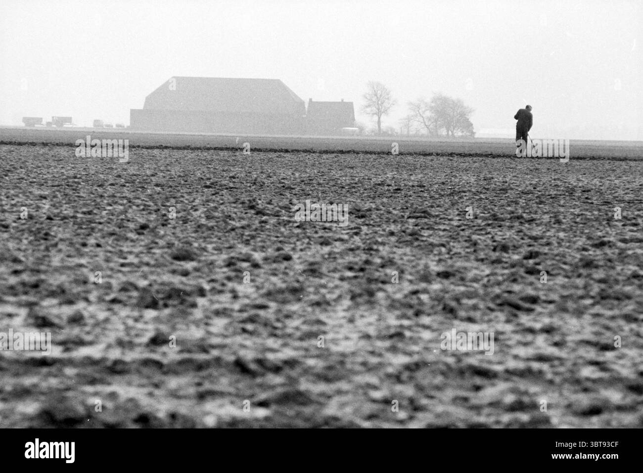 Hoofddorp N/V agricoltore al lavoro. Agriculture Hoofddorp Paesi Bassi, Whizgle News, Dutch Desk, Paesi Bassi, 1950 - 2000 il 13-03-1990. Questi argomenti sono mostrati nell'immagine. La scena è ambientata in un vasto paesaggio agricolo, caratterizzato da una tavolozza monocromatica che si appoggia pesantemente su sfumature di grigio e nero. Il primo piano è caratterizzato da un terreno coltivato con toni terrosi, dove il terreno è grezzo e irregolare, rivelando i segni lasciati dagli attrezzi agricoli. Il terreno stesso sembra incrinato e asciutto, suggerendo un'area arida o arata di recente. A metà strada si trova una figura solitaria, Foto Stock