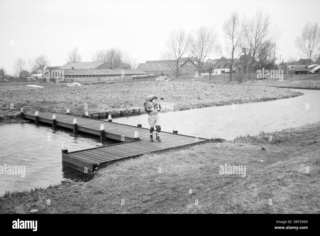 Sig. Ahlers Walker Ridderkerk Walking Ridderkerk, Whizgle News, Dutch Desk, Paesi Bassi, 1950 - 2000 il 09-04-1986. L'immagine mostra questi argomenti. La scena cattura un momento tranquillo accanto a un corpo d'acqua, dove una stretta passerella in legno conduce fuori sulla superficie dell'acqua. Il sentiero è fiancheggiato da piccoli pali bianchi che ne esaltano il fascino rustico. In primo piano, una figura cammina lungo la passerella, portando un oggetto sulla schiena, suggerendo un senso di scopo o di avventura. L'ambientazione presenta una tavolozza delicata e silenziosa dominata da sfumature di grigio e tonalità di terra morbide, che trasmettono un'atmosfera serena e allo stesso tempo discreta Foto Stock
