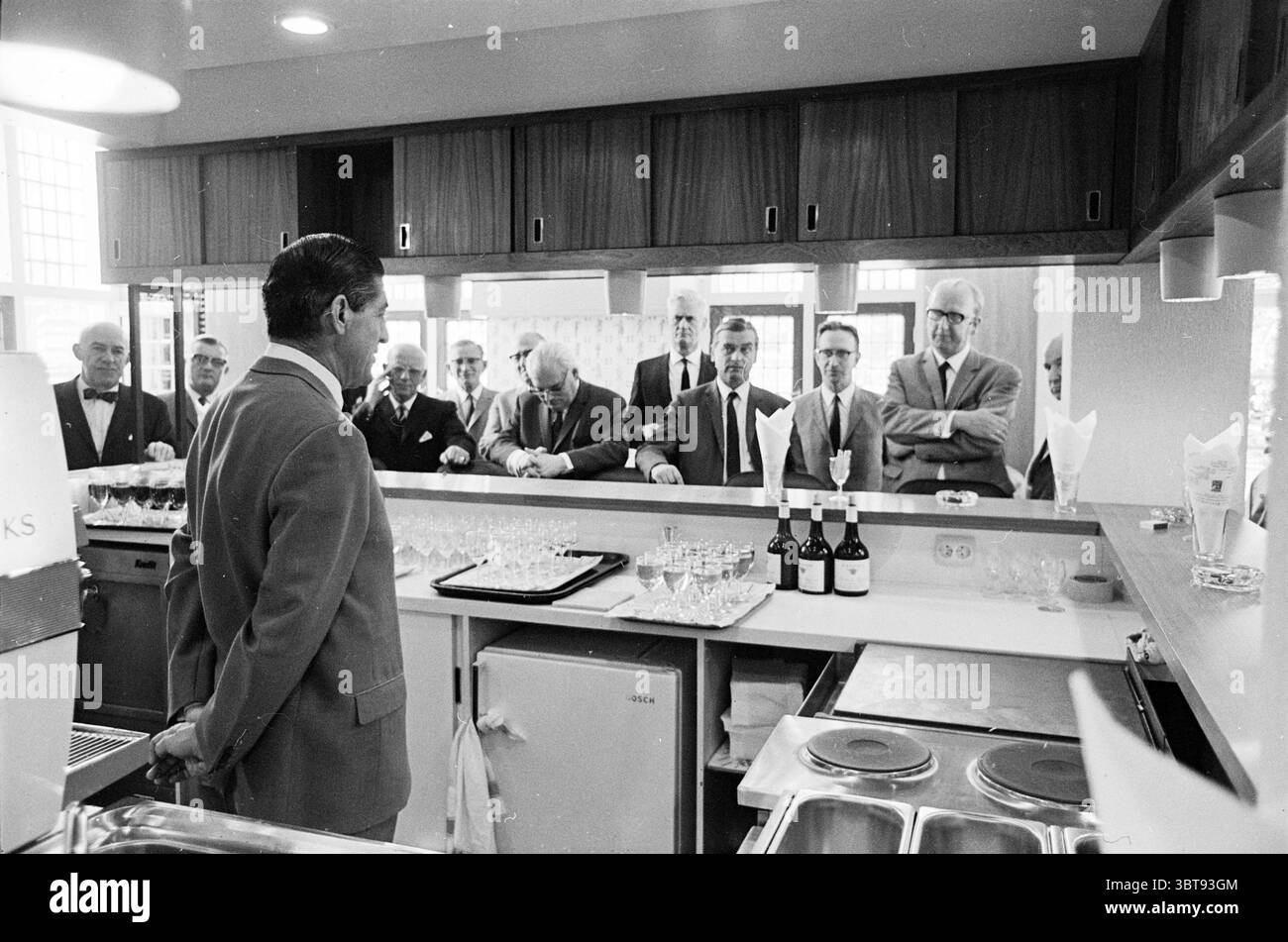 Apertura della caffetteria NZH Amsterdam North South Hollandse Vervoersmaatschappij N.V. NZ, Whizgle News, Dutch Desk, Paesi Bassi, 1950 - 2000 il 31-05-1968. L'immagine mostra questi argomenti. La scena è ambientata in una calda e invitante cucina con un ripiano lucido rivestito con varie attrezzature da cucina, tra cui un lavandino scintillante e pentole da cucina in metallo. In primo piano, un uomo sta con le spalle allo spettatore, vestito con una tuta formale con una distinta giacca di colore chiaro, trasudando un'aria di autorità e fascino. È impegnato con un pubblico invisibile, probabilmente introducendo un Foto Stock