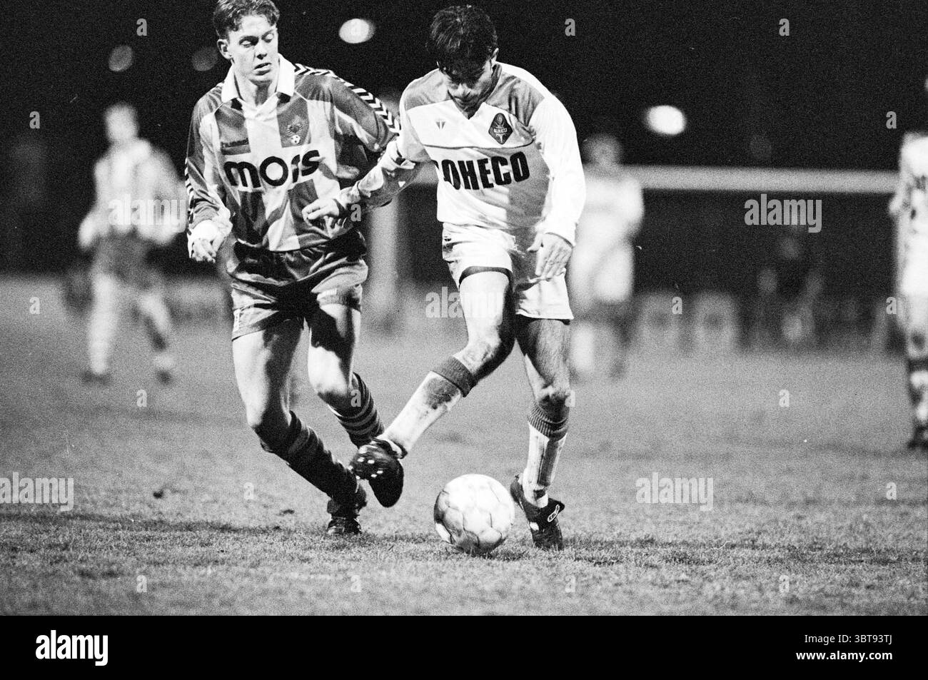 Telstar - Go Ahead Eagles, Whizgle News, Dutch Desk, Paesi Bassi, 1950 - 2000 on 27-03-1992. L'immagine mostra questi argomenti. La scena raffigura un momento dinamico in una partita di calcio sotto un'illuminazione artificiale mirata, che crea un'atmosfera suggestiva. Due giocatori sono il punto focale, entrambi con divise distinte: Uno in maglia bianca con dettagli arancioni e l'altro in un abito a righe rosse e bianche. Il giocatore con la maglia bianca sta facendo dribbling attivo sulla palla, dimostrando acuta concentrazione e agilità durante le manovre in campo. La sua postura suggerisce una miscela di determinazione e finezza, Foto Stock