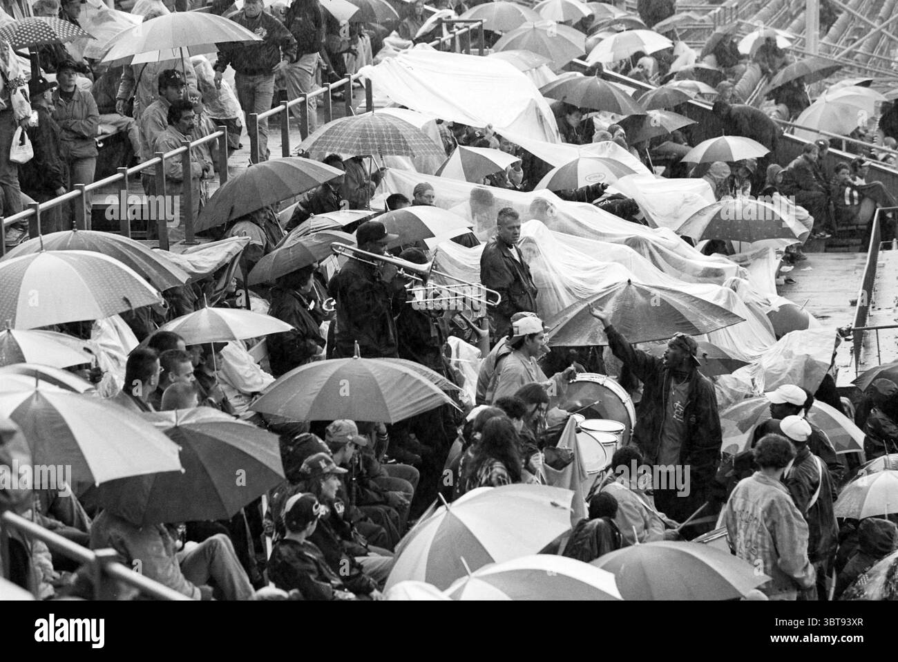 HHW Rain [Haarlem Baseball Week], Whizgle News, Dutch Desk, Paesi Bassi, 1950 - 2000 on 28-06-1996. Questi sono gli elementi dell'immagine. La scena raffigura un ambiente all'aperto affollato, probabilmente uno stadio, pieno di un gruppo diversificato di persone riunite sotto un cielo spaventoso e coperto. Numerosi ombrelli creano un vivace arazzo di colori, con rossi, blu, gialli e verdi luminosi che contrastano con l'ambiente grigio tenue. Alcuni ombrelli vengono ribaltati all'interno verso l'esterno o presentano gocce di pioggia che cadono sulle loro superfici, a indicare condizioni meteorologiche avverse. I partecipanti sono vestiti con un mix di casua Foto Stock