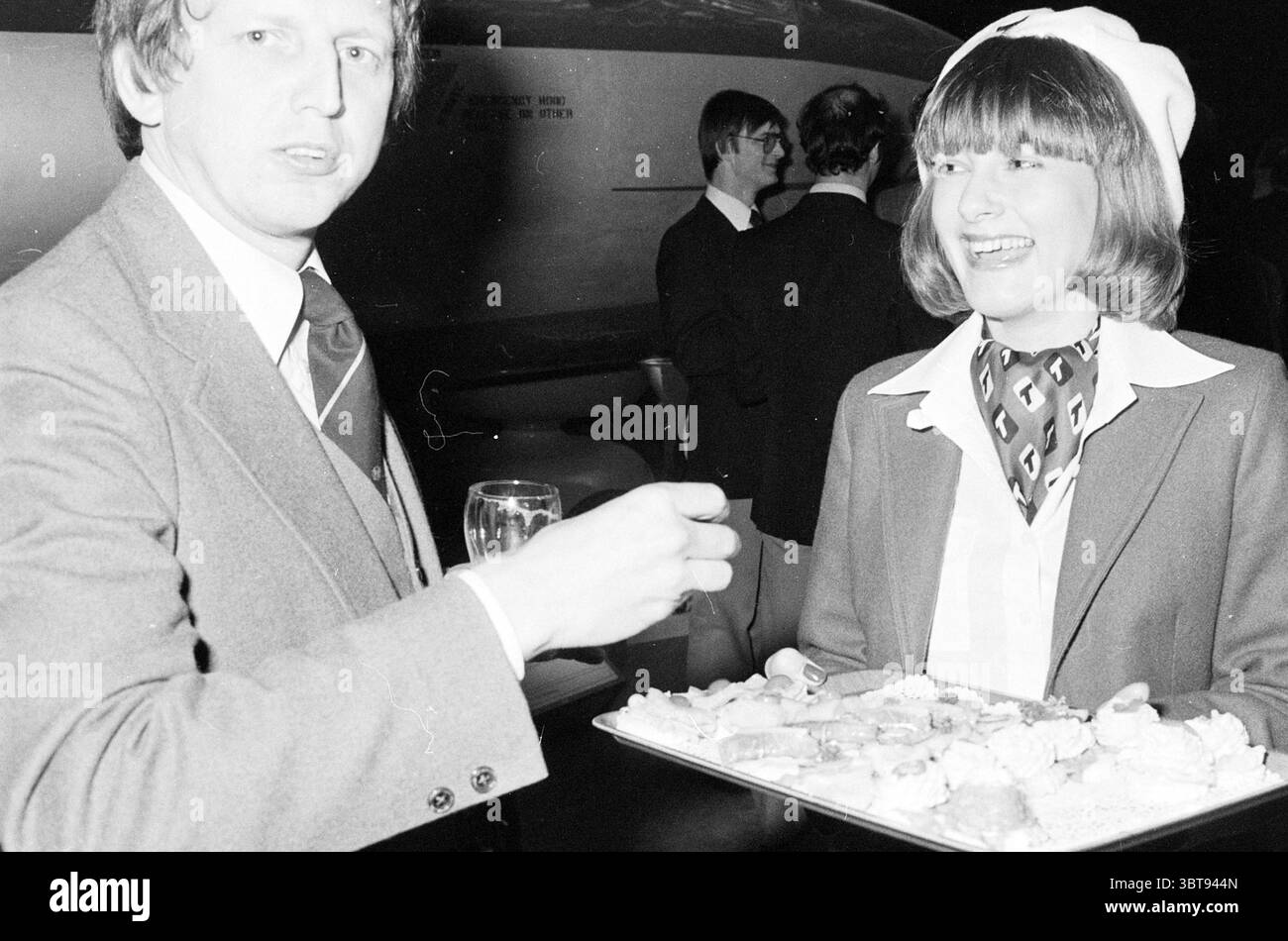 Ricevimento Transavia in una stanza con vecchi aerei e modelli di aerei, Whizgle News, Dutch Desk, Paesi Bassi, 1950 - 2000 il 10-03-1979. Questi sono gli elementi dell'immagine. La scena cattura un vivace raduno al coperto con due figure ben esposte. A sinistra, un uomo con una tuta si impegna con lo spettatore, tenendo in mano un vetro che riflette la luce ambientale. La sua acconciatura è ordinata e mostra un'espressione che combina attenzione e curiosità. A destra, una donna tiene in mano un vassoio d'argento pieno di un assortimento di spuntini colorati, che mostrano una varietà di forme e texture. S Foto Stock
